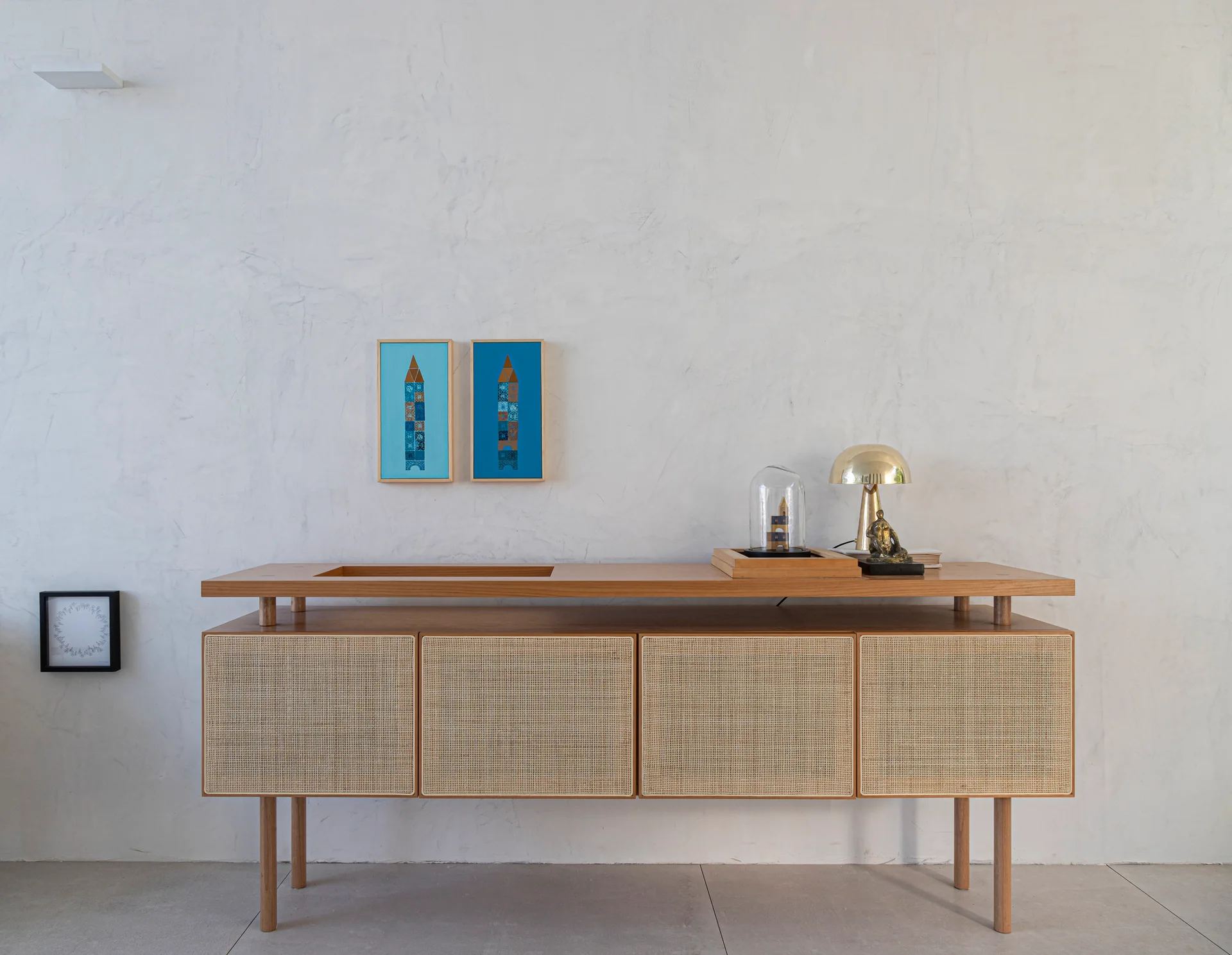 A wooden sideboard with four cane mesh doors stands against a textured white plaster wall beneath two framed blue art pieces.