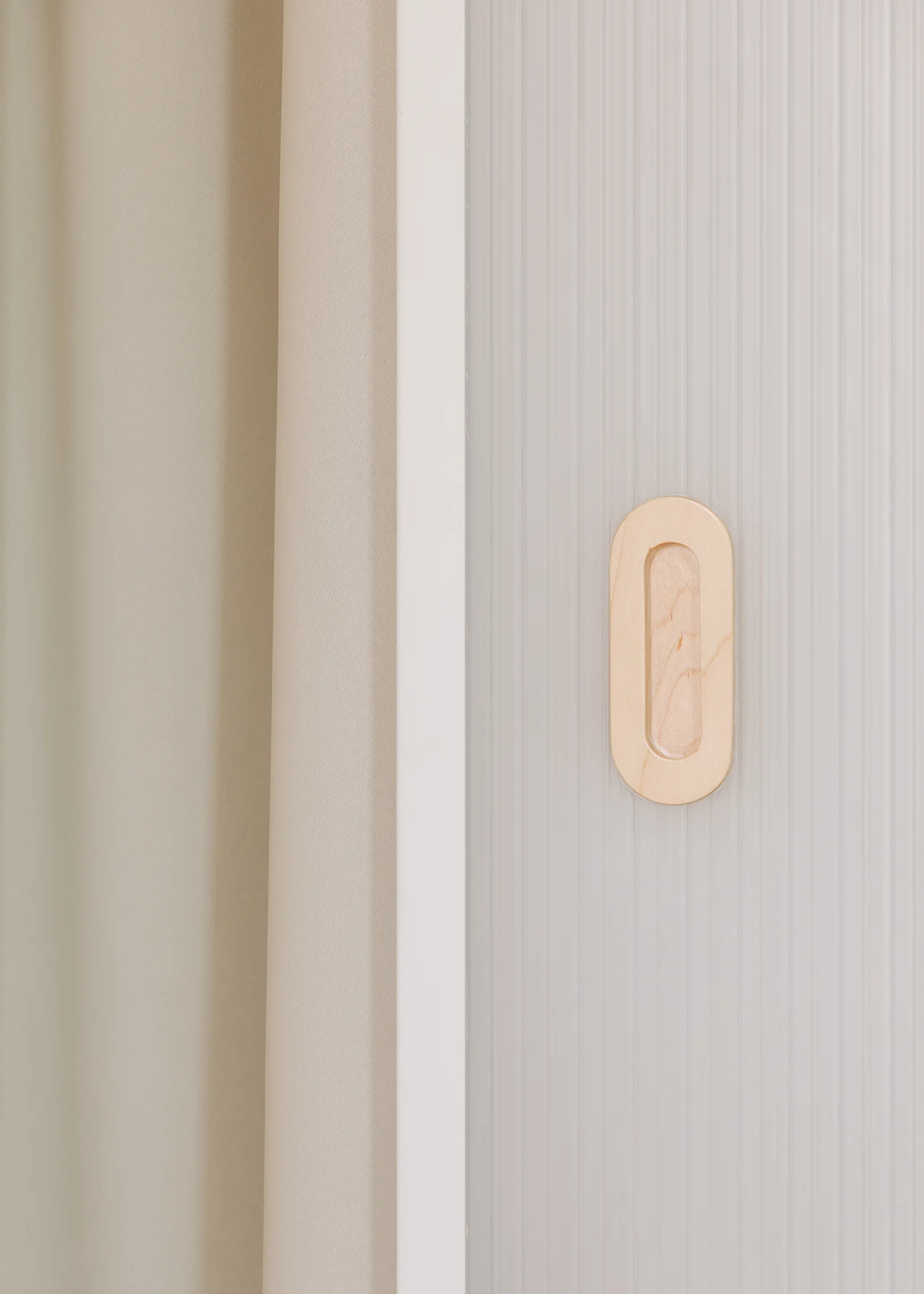 A close-up of a pale, vertically ribbed door panel with a recessed oval wooden handle next to a beige fabric curtain.