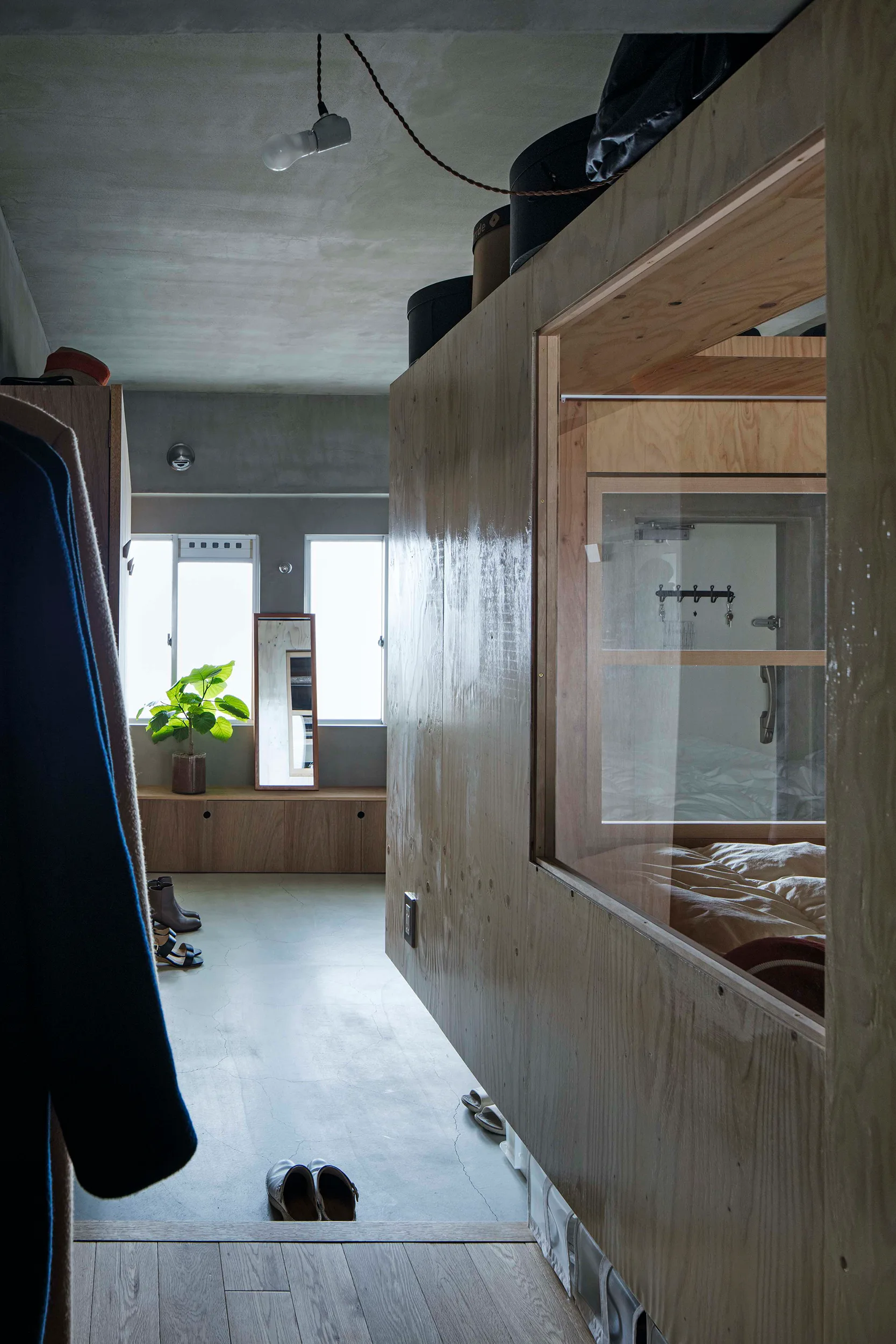 A minimalist studio apartment featuring a large plywood partition wall with a glass window enclosing a bed, set against grey concrete floors and ceiling.