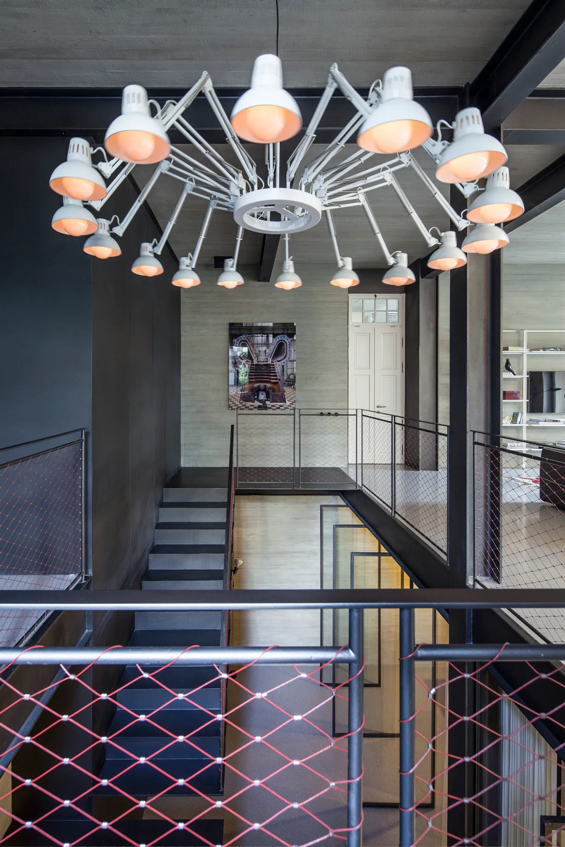 A multi-level industrial staircase with black metal steps, wire mesh railings, and a large white multi-arm articulating lamp chandelier hanging above.