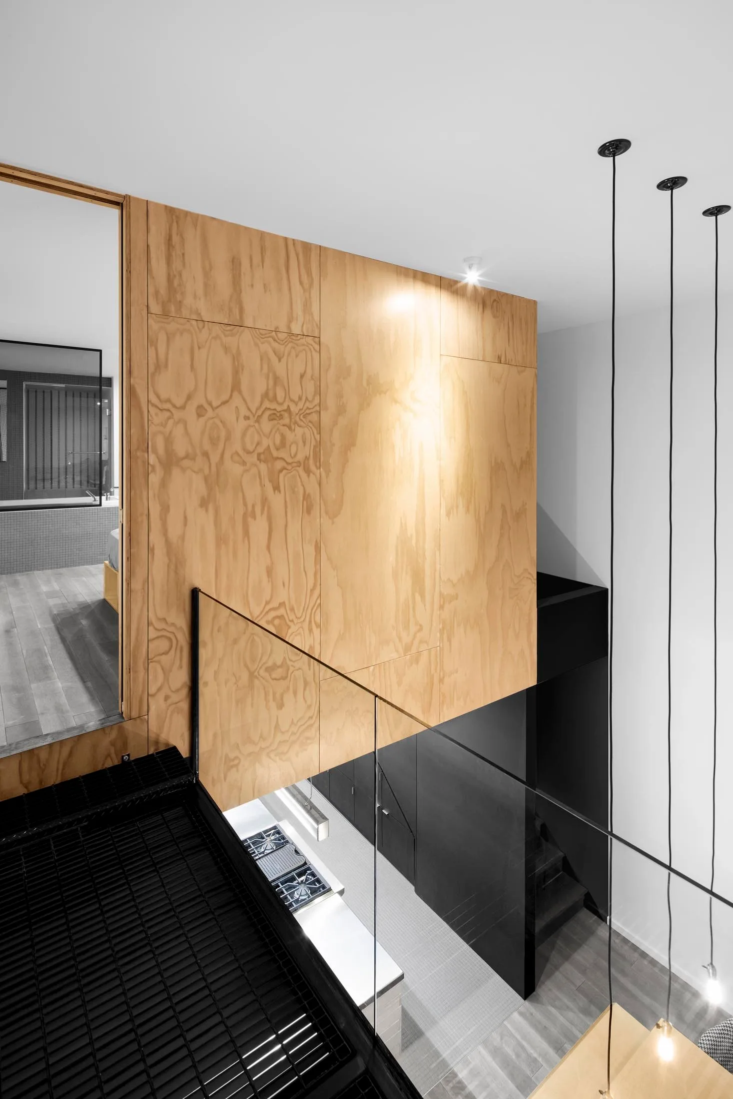A high-angle view of a modern staircase area featuring large plywood wall panels, a clear glass railing, and a black gridded metal landing.
