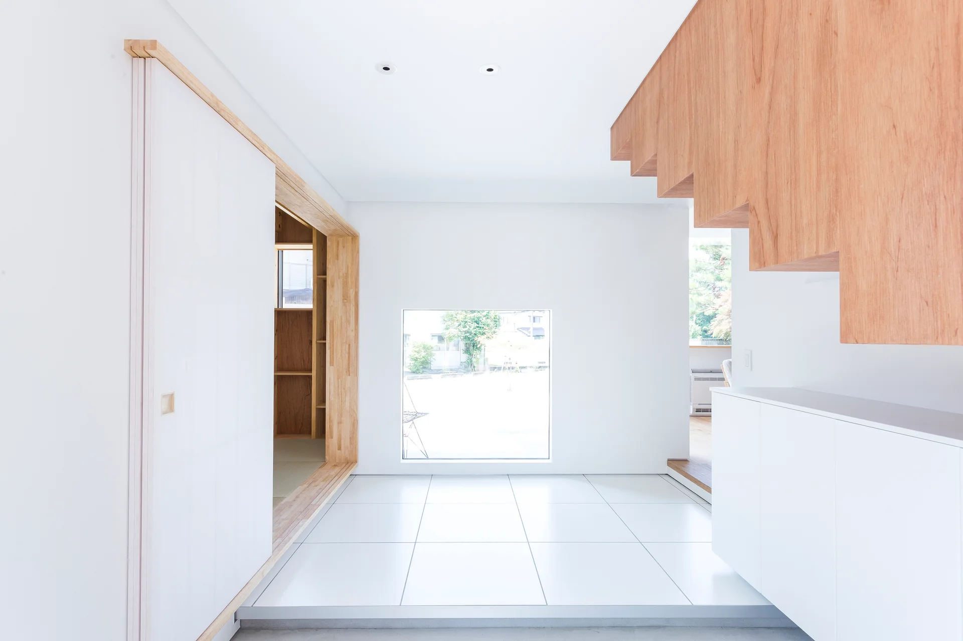 A minimalist white entryway featuring a suspended wooden staircase, a square window, white floor tiles, and a wood-trimmed doorway.
