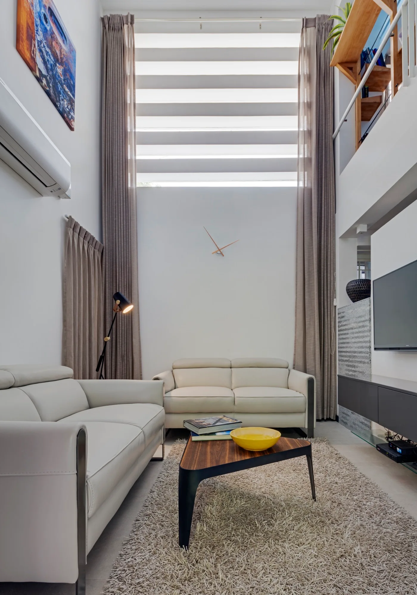 A double-height living room features light cream leather sofas, a triangular wood coffee table on a shag rug, and tall beige drapes.