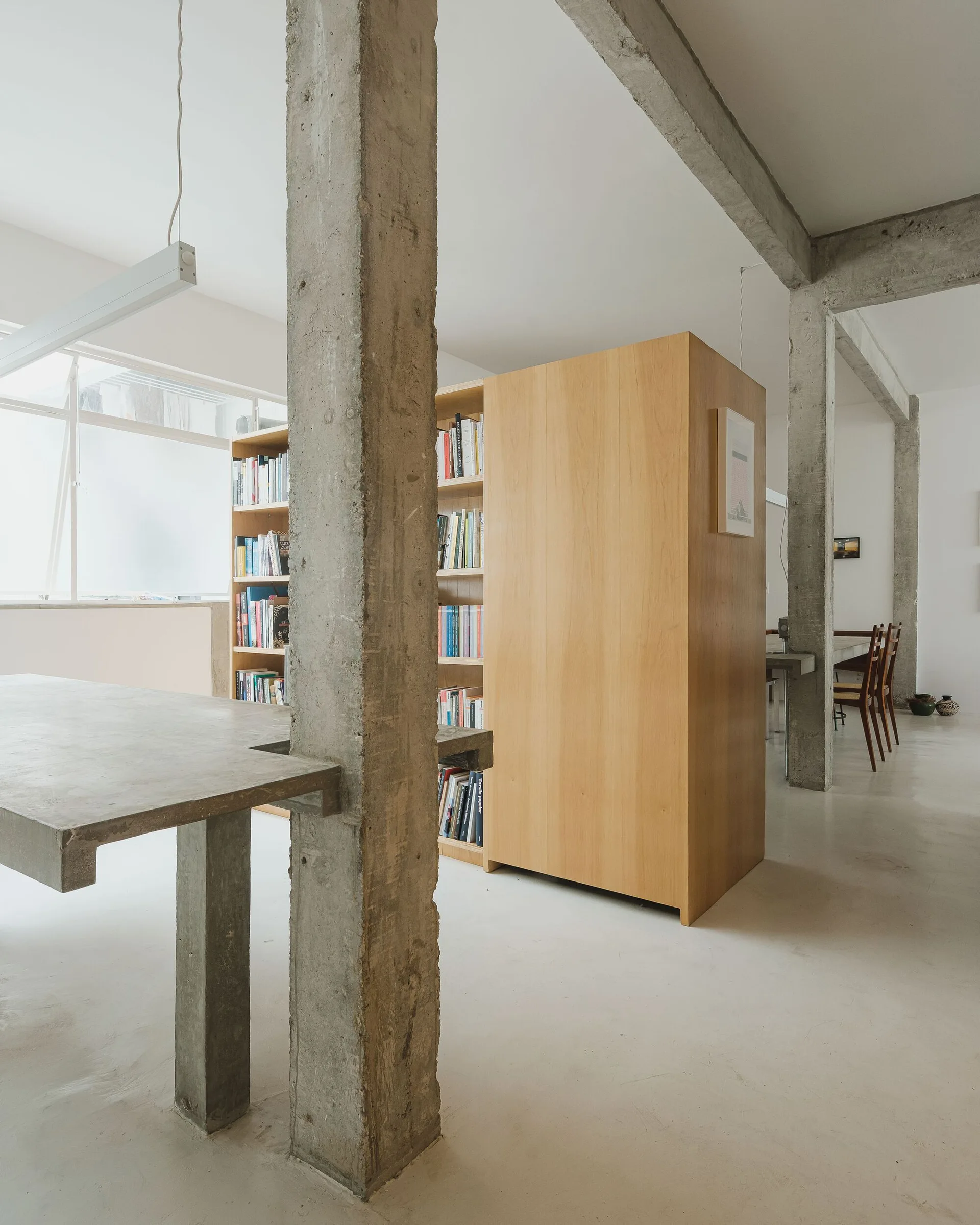 A modern loft interior featuring rough exposed concrete columns, a light wood double-sided bookcase, and a concrete dining table.