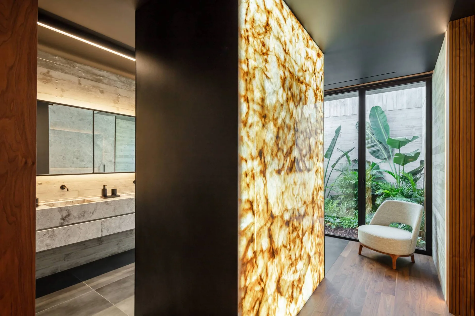 A modern bathroom features a floating marble vanity, a backlit glowing stone partition wall, and a lounge chair facing a glass wall with garden views.