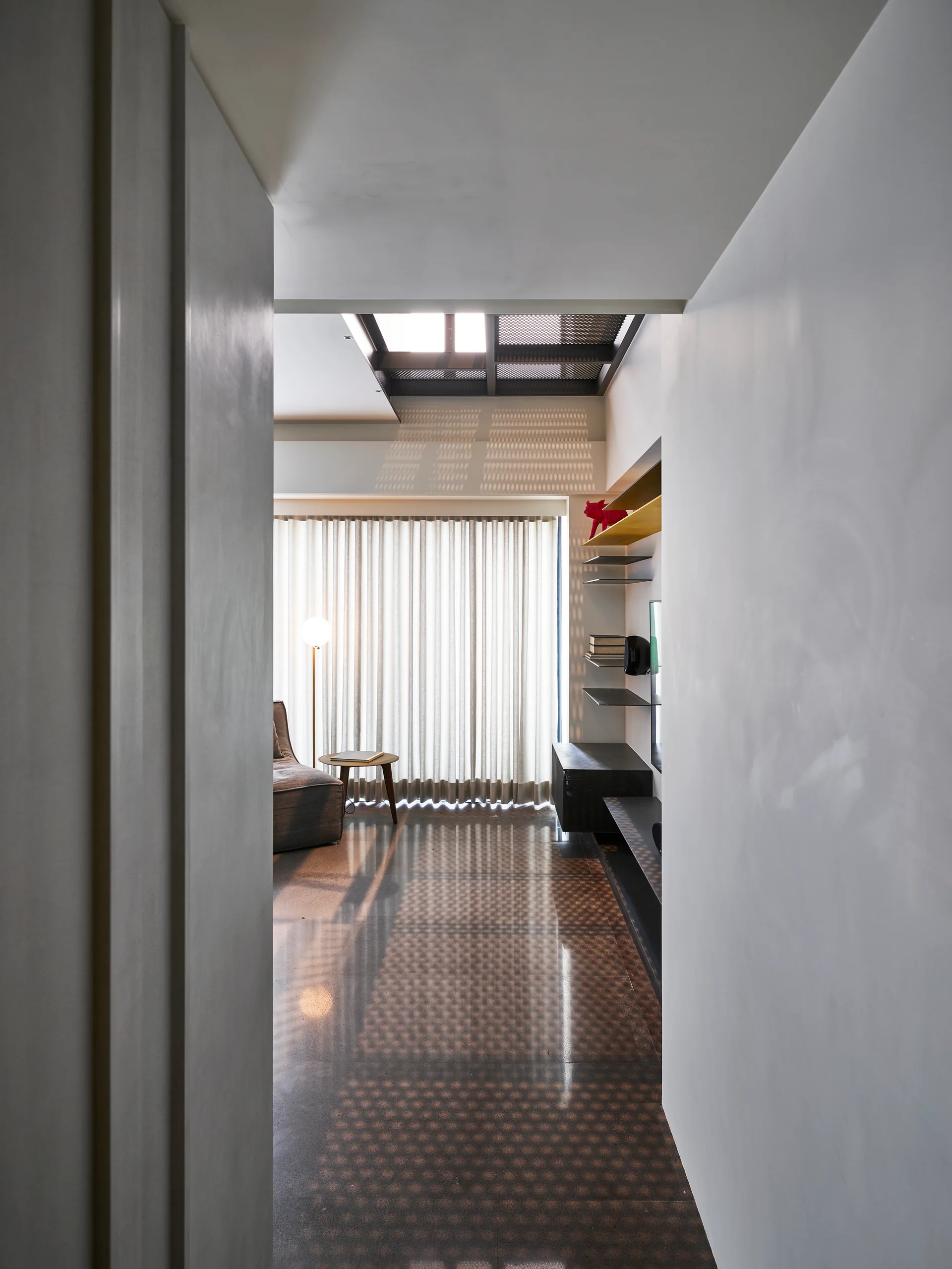 A minimalist living room viewed from a narrow hallway, featuring a floor-to-ceiling curtain, a grey armchair, and geometric shadows from a skylight.