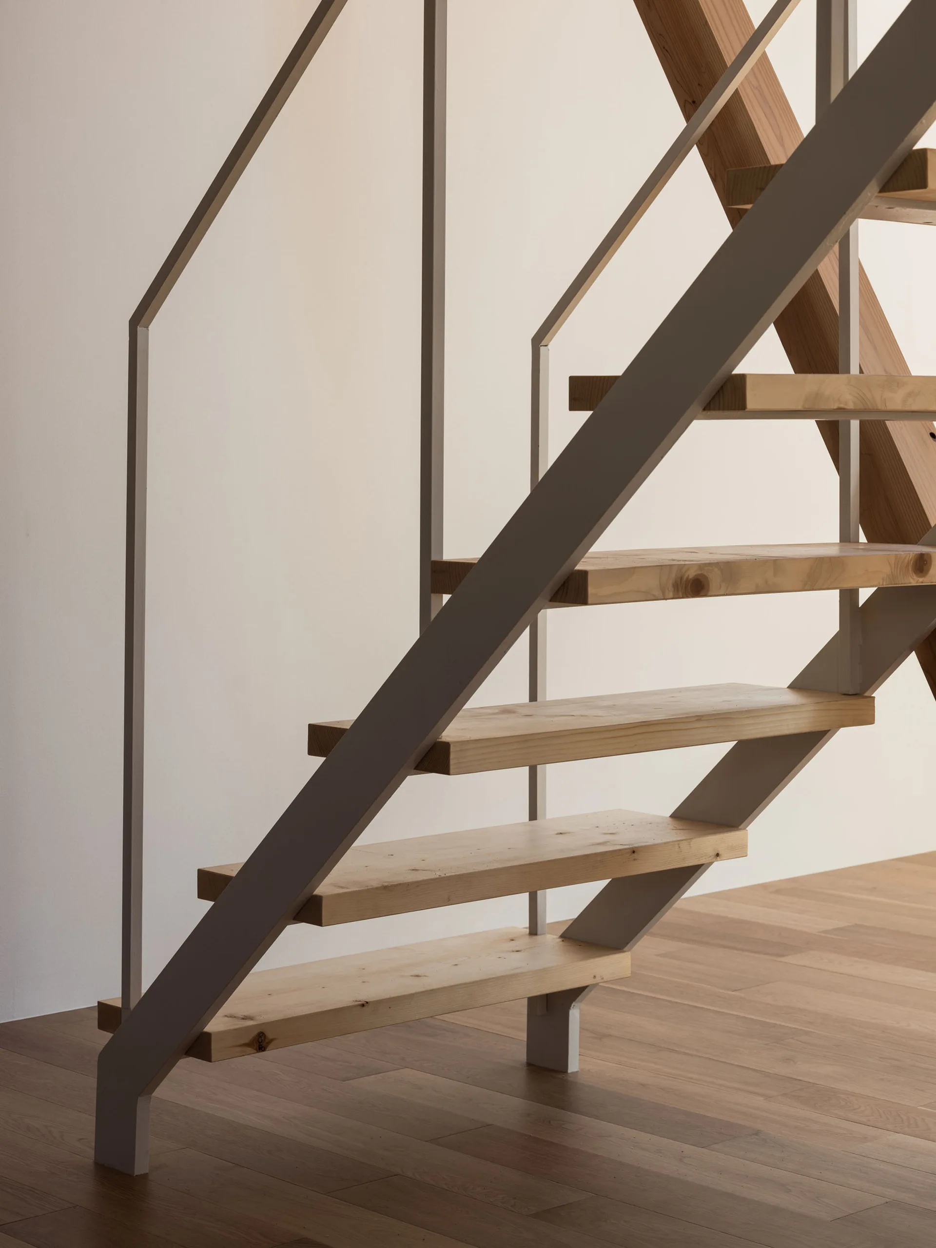 A minimalist open-riser staircase features light wood treads supported by a slim metal frame against a plain off-white wall.