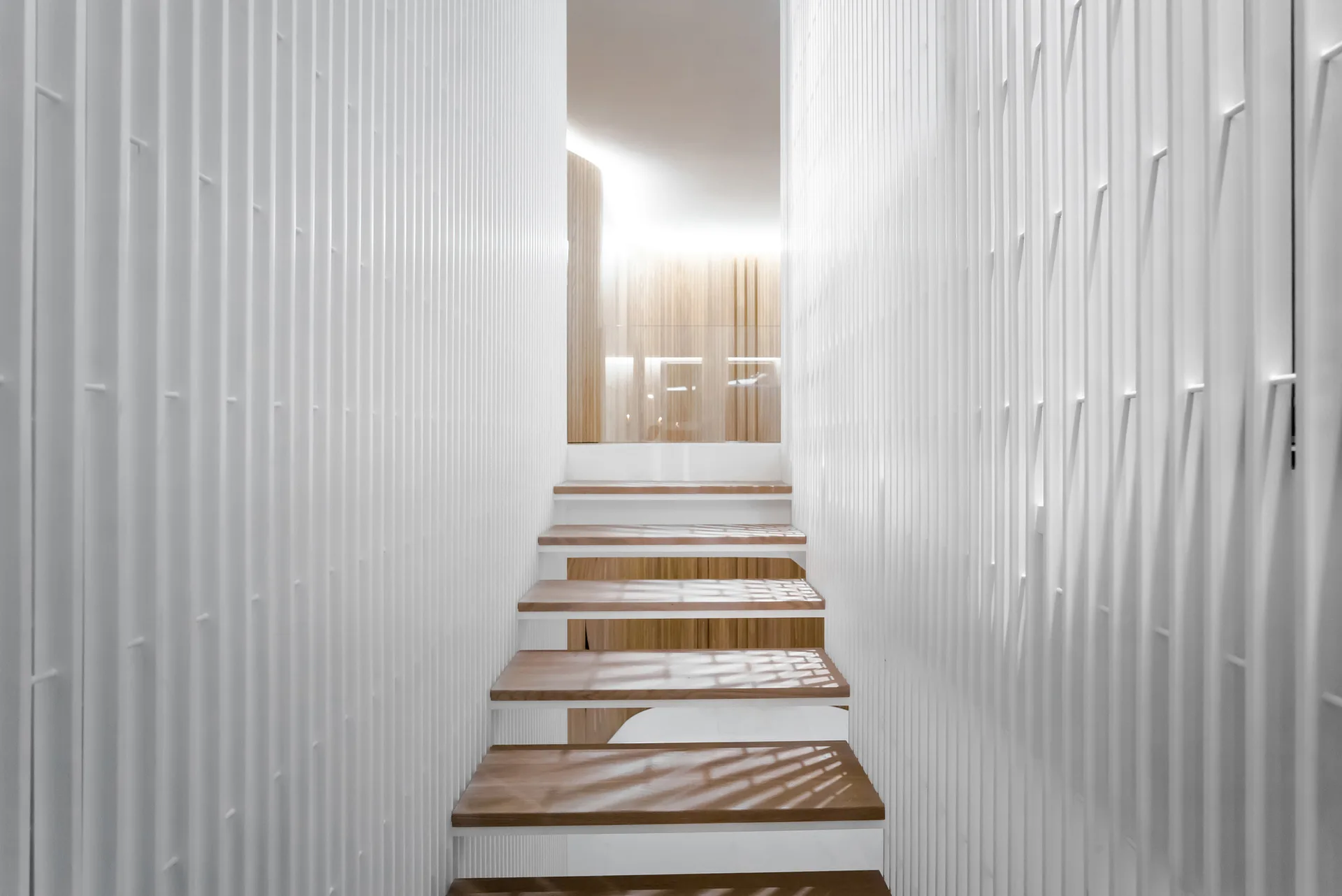 A modern staircase featuring light wood treads flanked on both sides by floor-to-ceiling white vertical slatted wall panels.