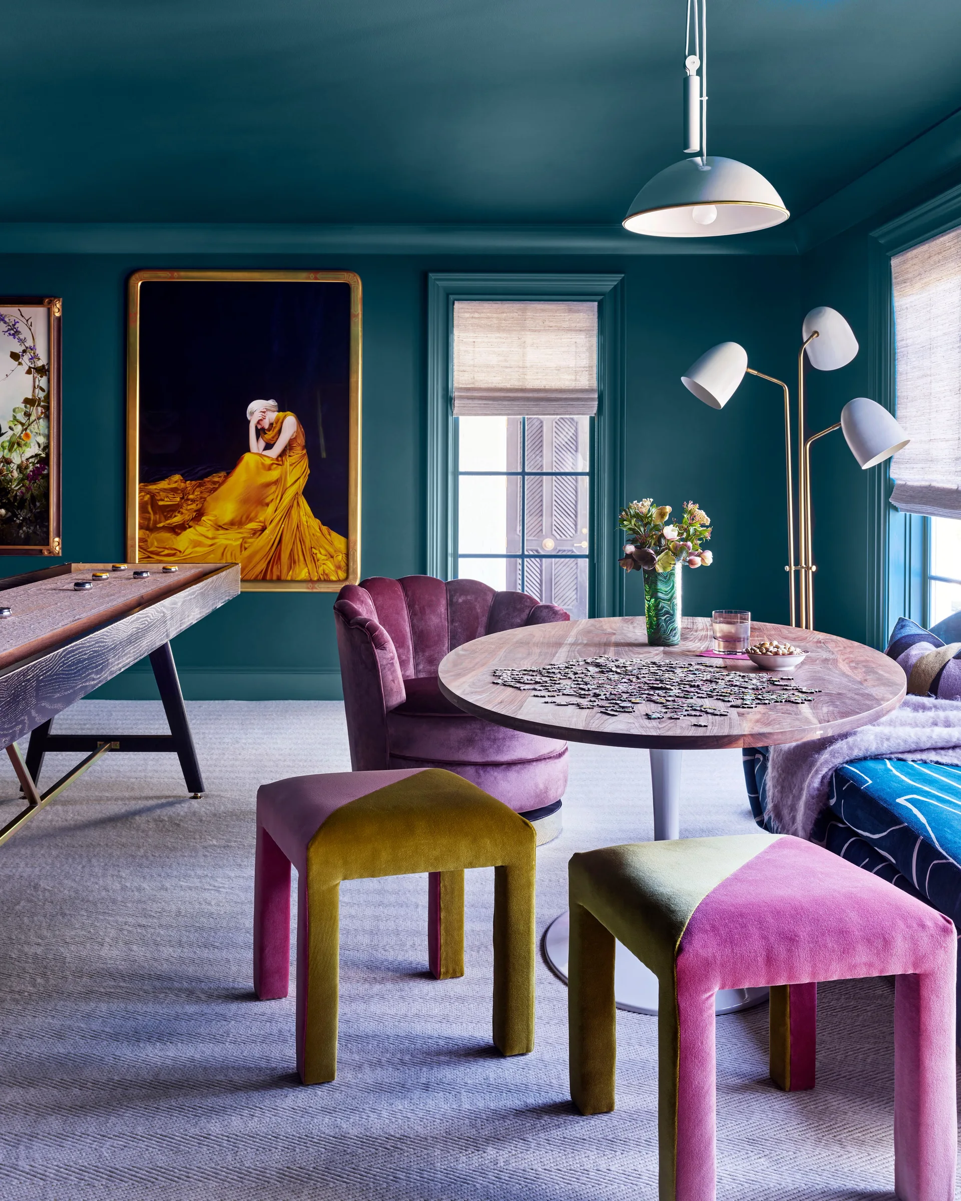A game room with deep teal walls and ceiling features a wooden shuffleboard table, a round dining table, and colorful velvet chairs and stools.