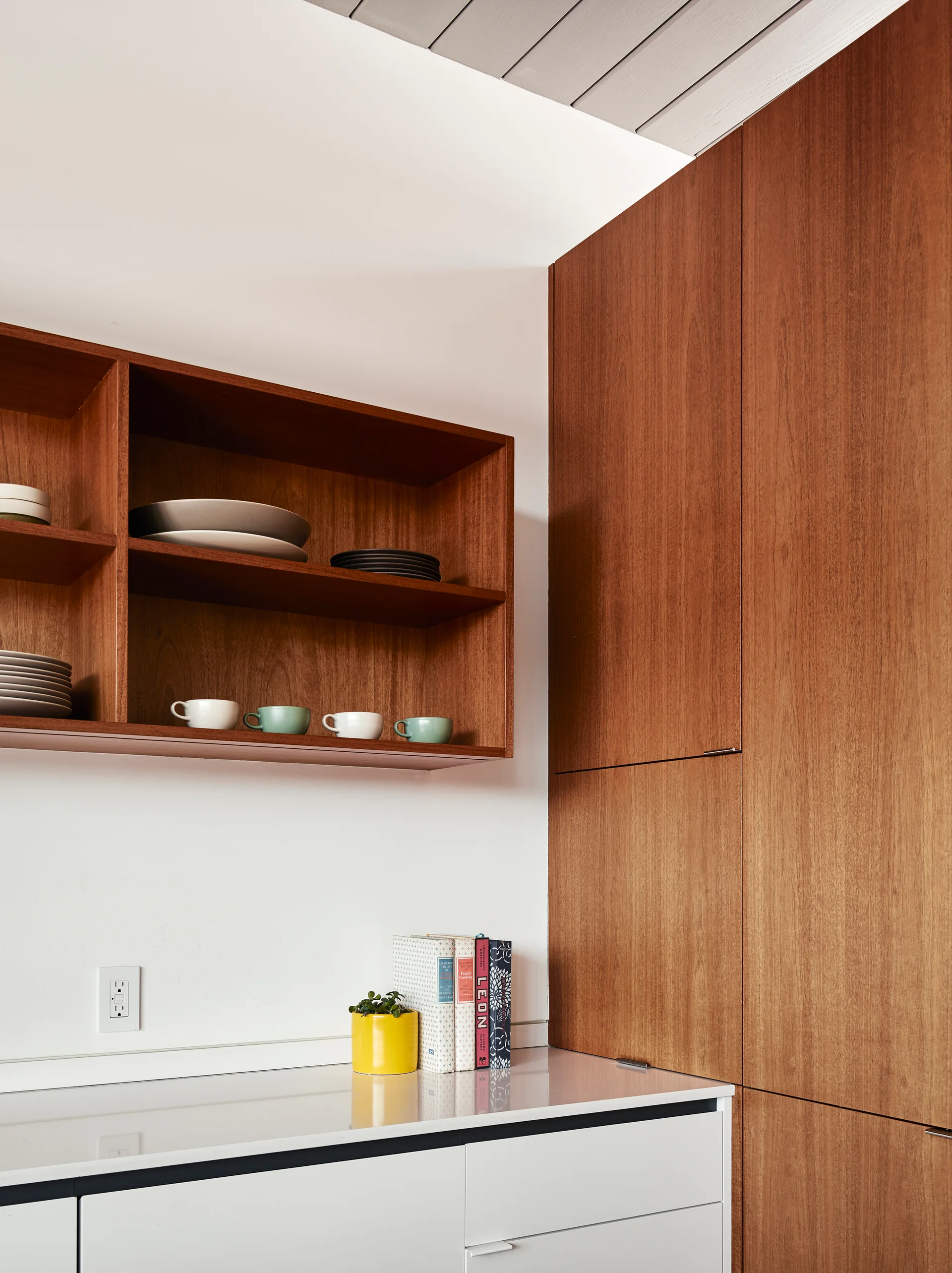 A modern kitchen corner featuring floor-to-ceiling wood grain cabinets, a white countertop, and matching open wood shelving with stacked dishes.