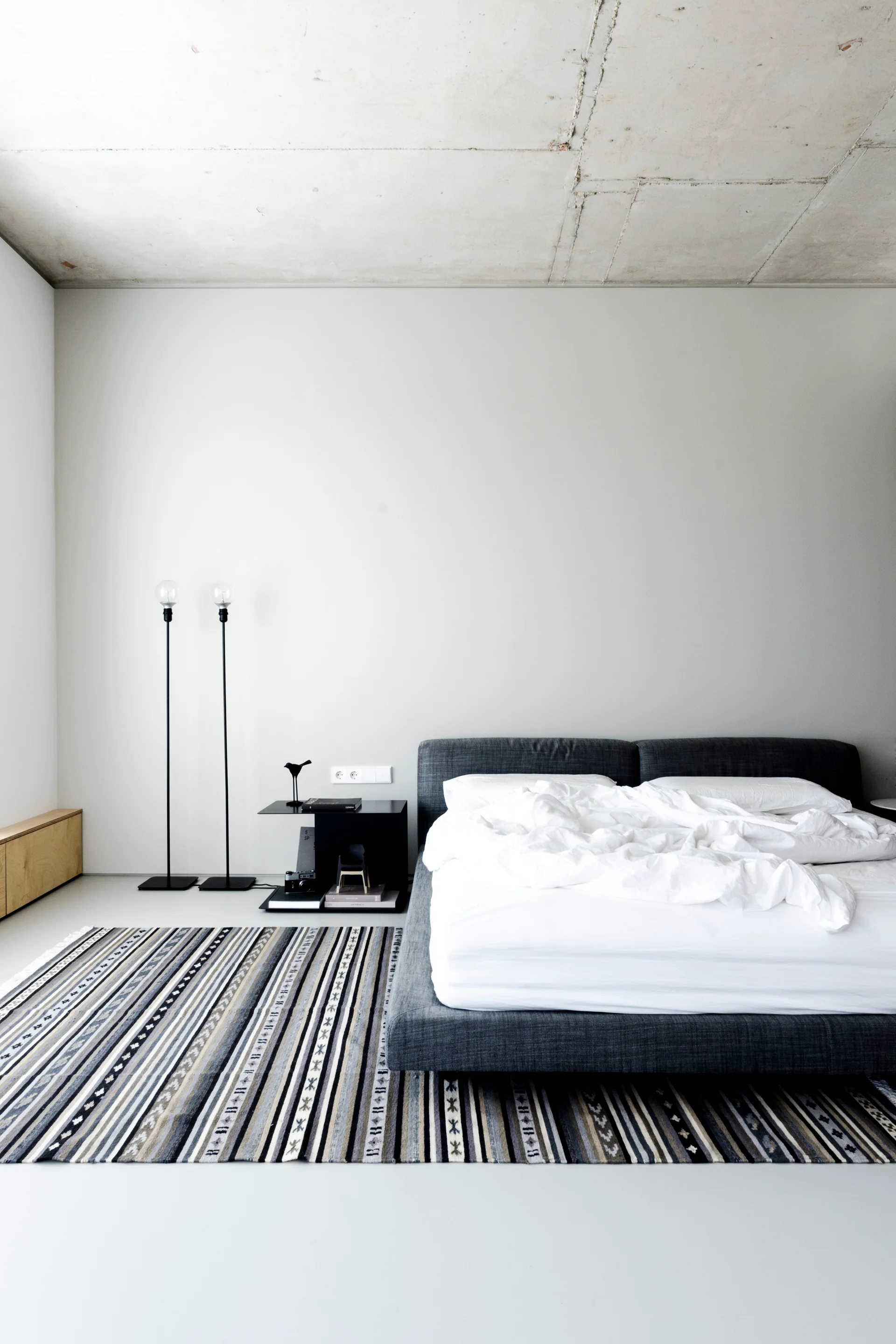 A minimalist bedroom featuring a charcoal grey upholstered bed and a multi-colored striped area rug beneath an exposed concrete ceiling.