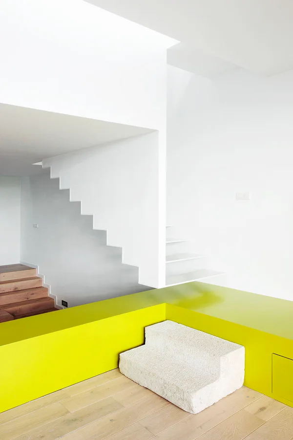 A minimalist white floating staircase ascends past a vibrant lime green raised platform with a rough textured stone step on light wood flooring.