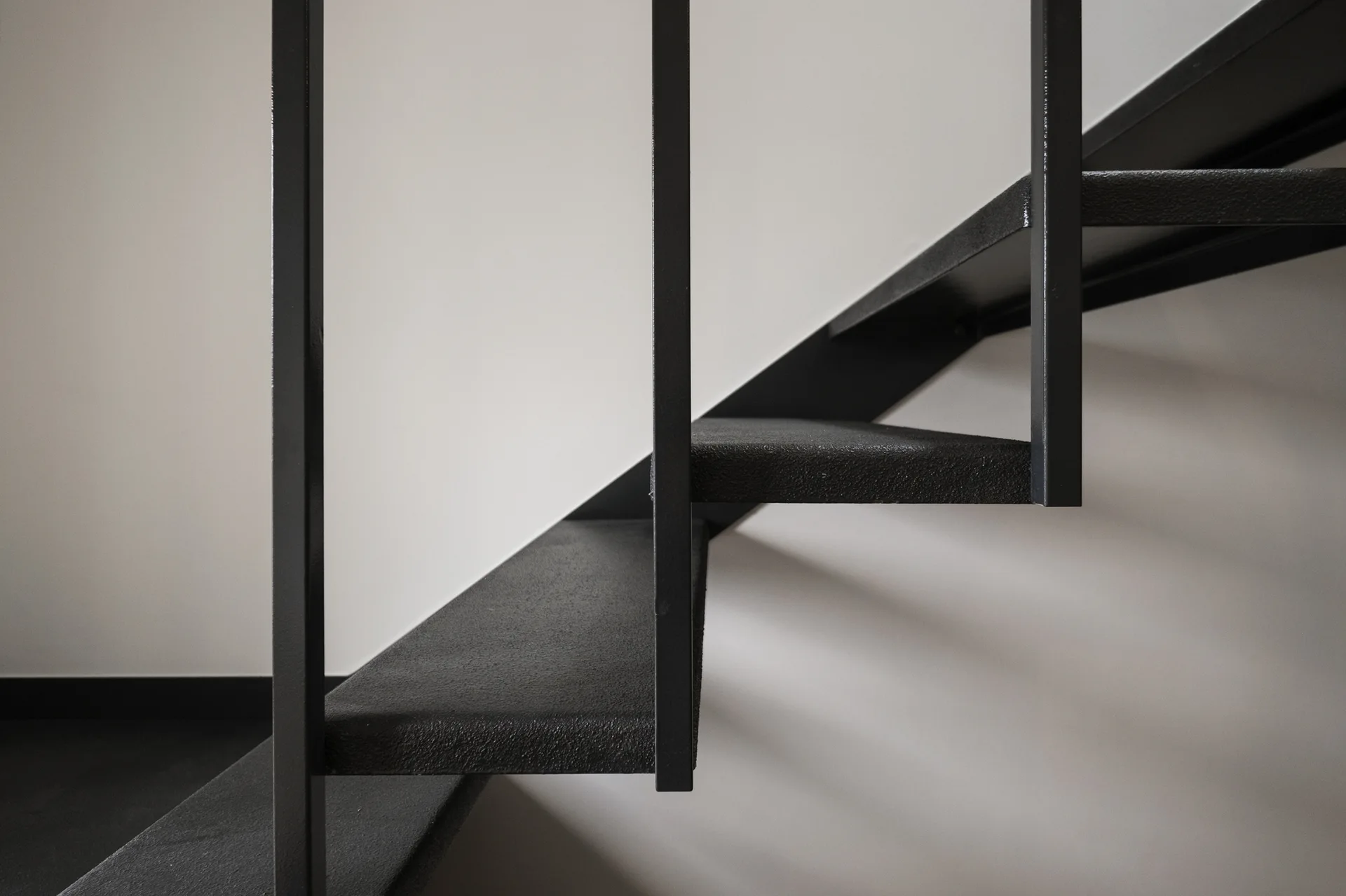 A close-up of a minimalist staircase featuring black textured metal open steps and vertical metal railing posts against a plain white wall.