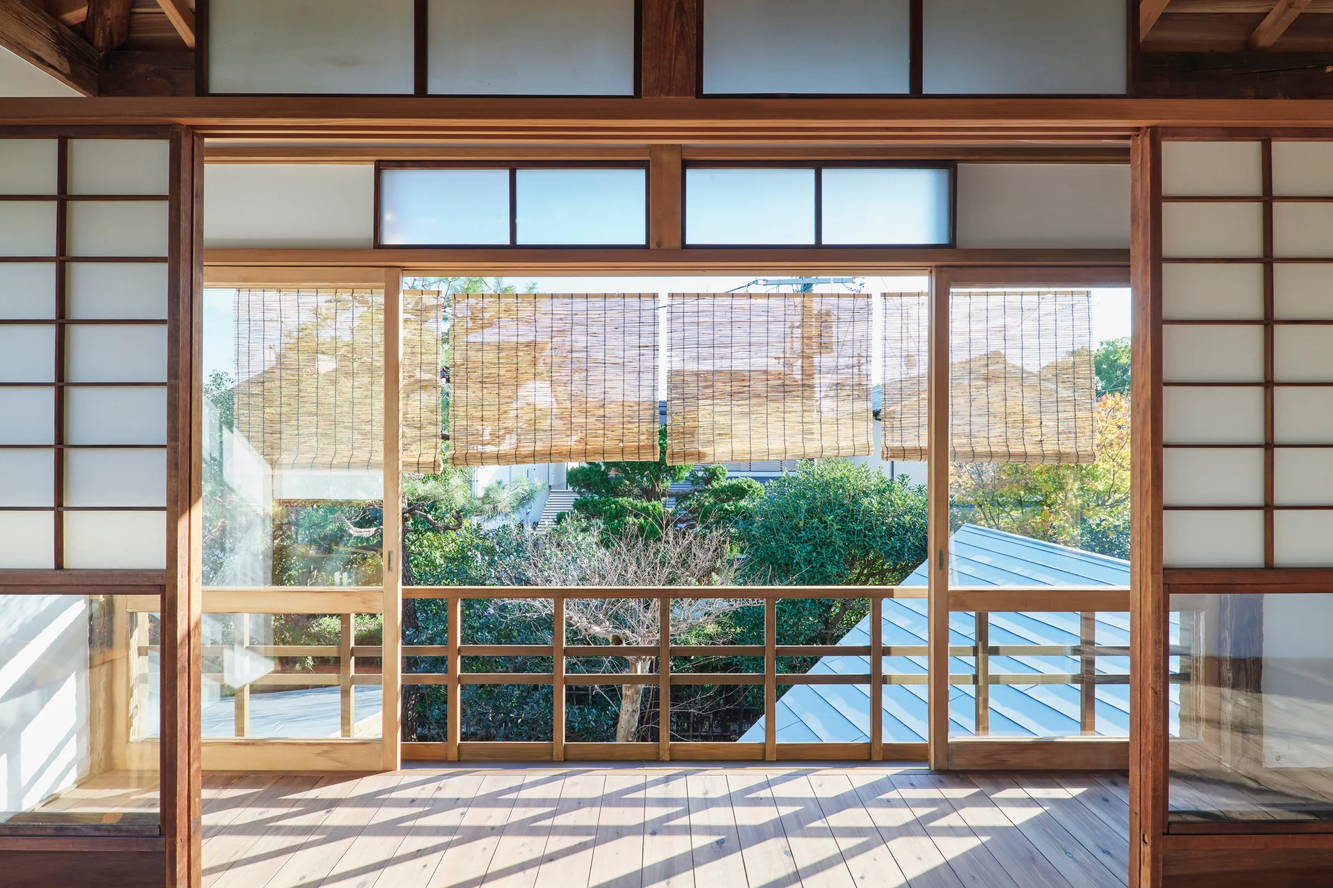A traditional Japanese room features wooden sliding shoji screen doors that open onto a deck with bamboo shades and a green garden view.