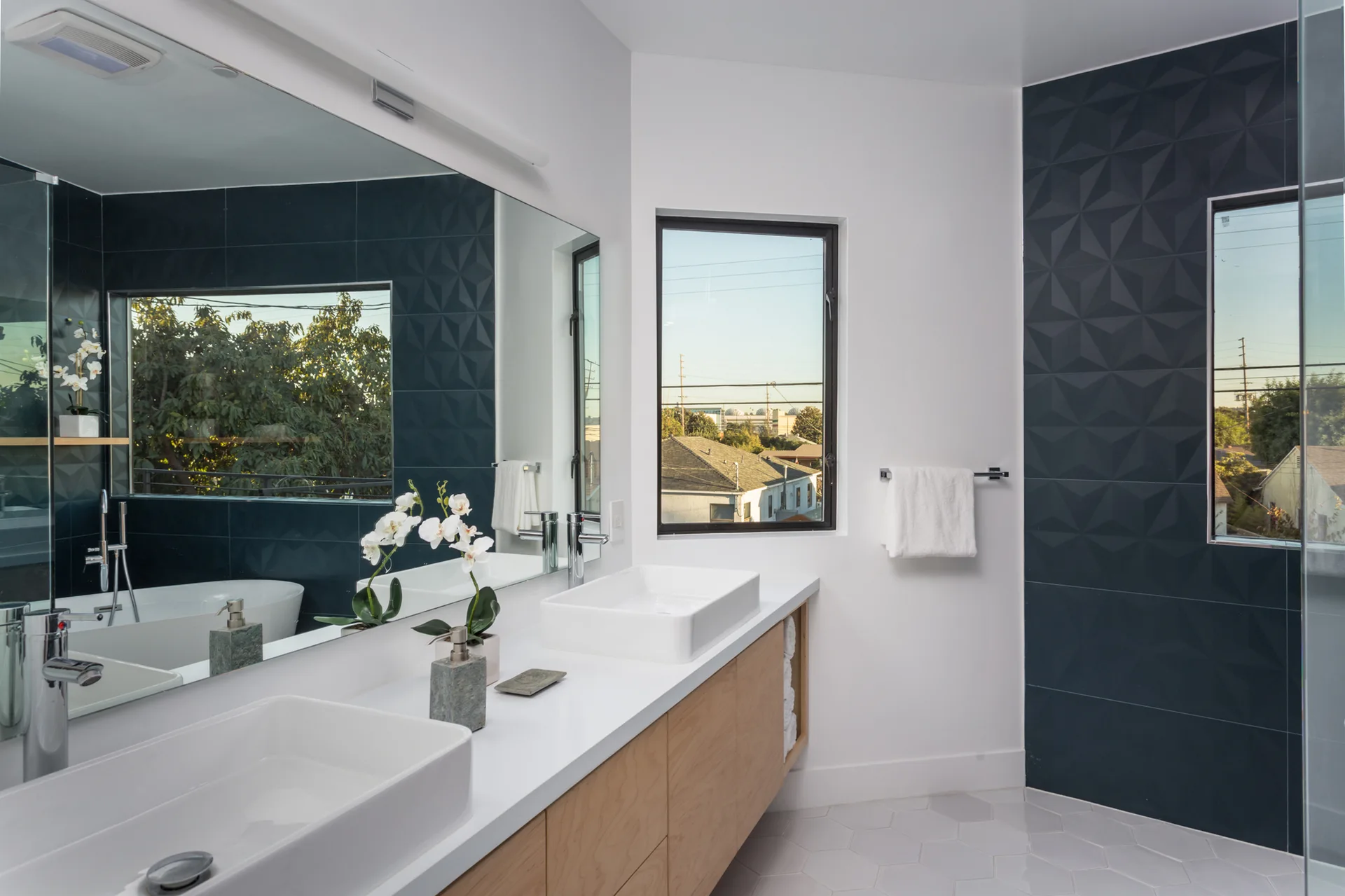 A modern bathroom featuring a light wood floating vanity with two white vessel sinks, a large mirror, and a dark blue geometric tile accent wall.
