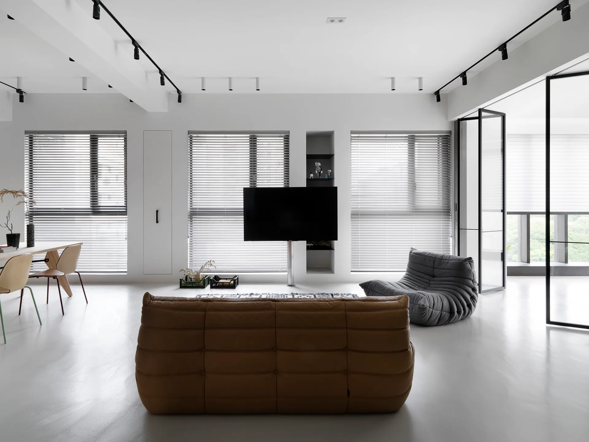A minimalist white living room featuring a tan tufted sofa, a grey lounge chair, and black track lighting with large windows in the background.