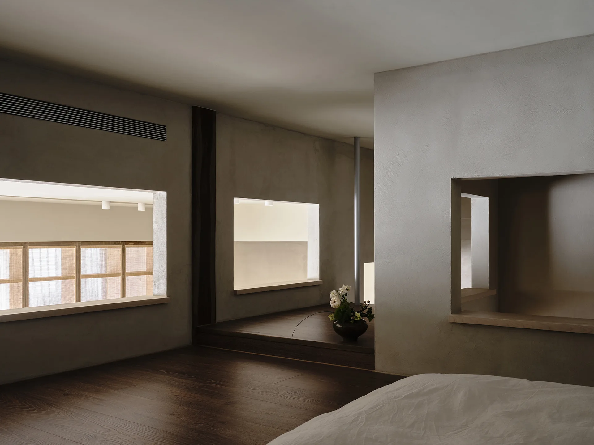 A minimalist bedroom featuring dark wood floors, gray textured plaster walls with rectangular cutouts, and a low platform transition.