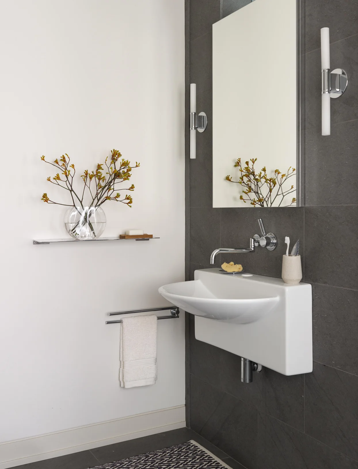 A modern bathroom features a white wall-mounted sink set against dark grey tiles, accented by a rectangular mirror, vertical tube sconces, and a white wall.