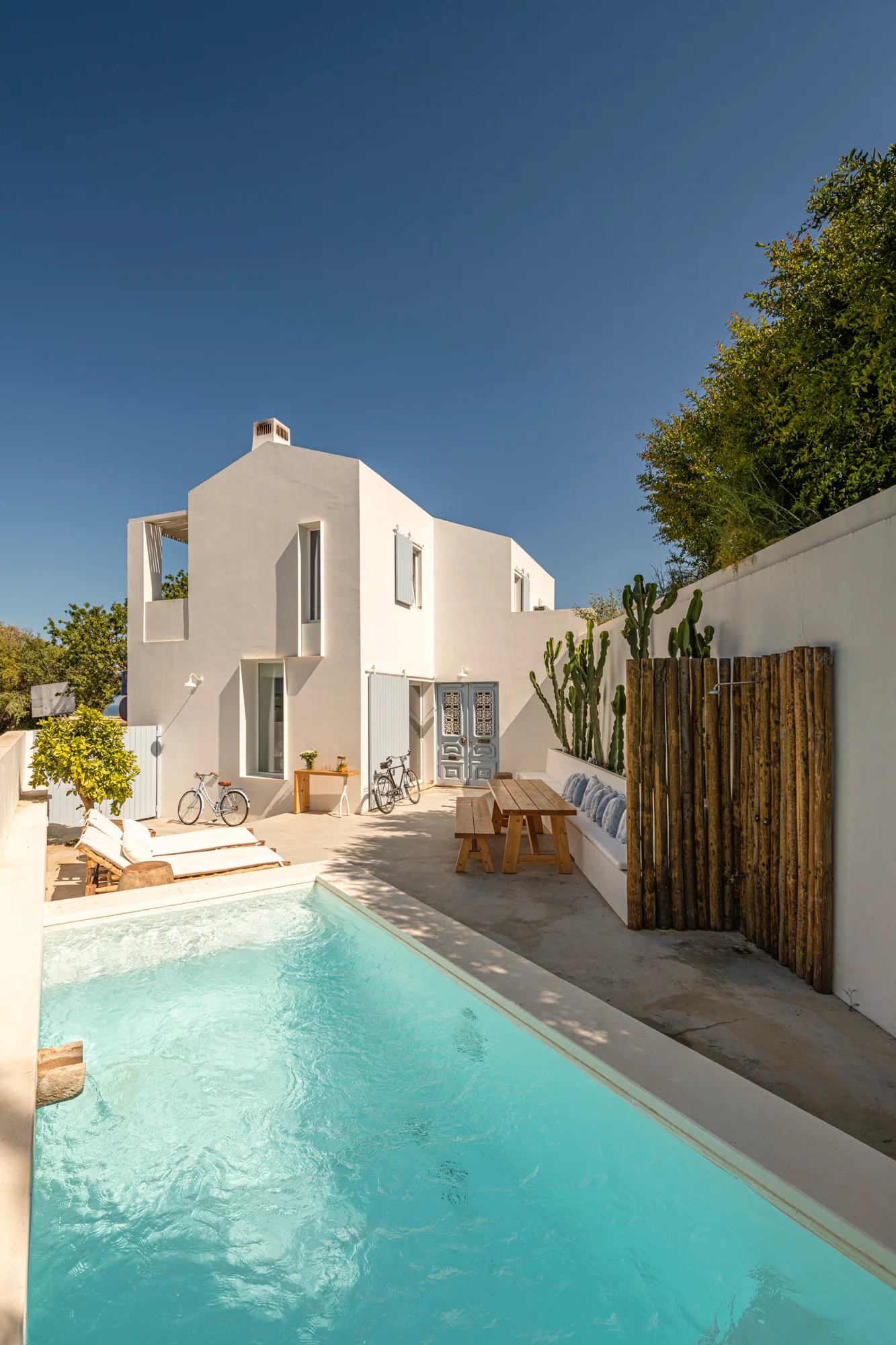 A rectangular swimming pool sits next to a white stucco house with a wooden dining table, built-in bench seating, and an outdoor shower.