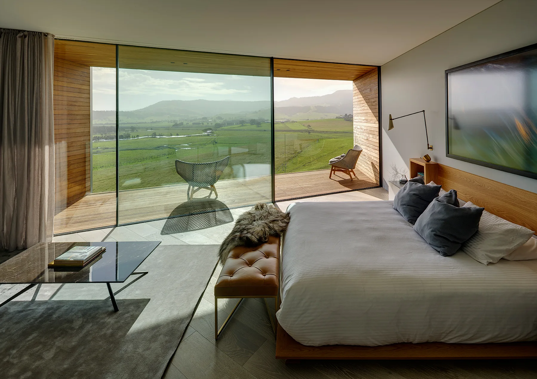 A modern bedroom features a low wood bed and a leather bench next to large sliding glass doors that open onto a wood-clad terrace.