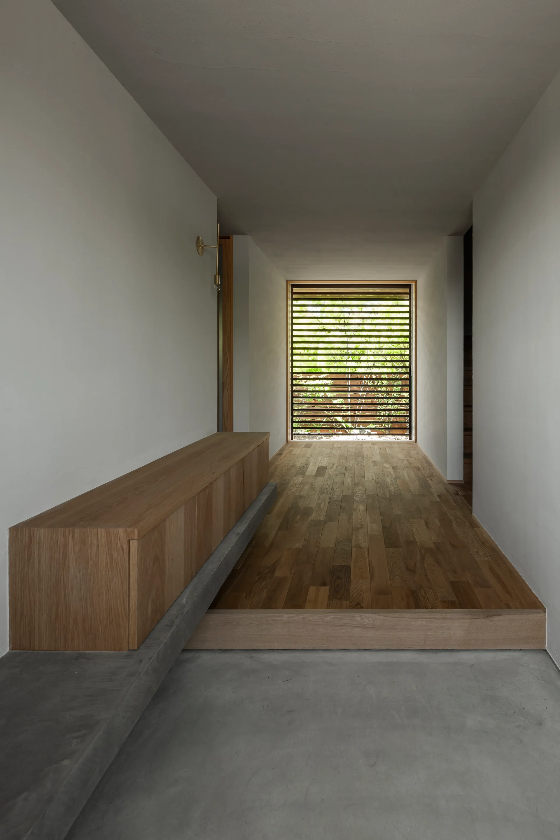 A minimalist hallway featuring a split-level floor with concrete and wood, a long built-in wooden bench, and a slatted window at the end.