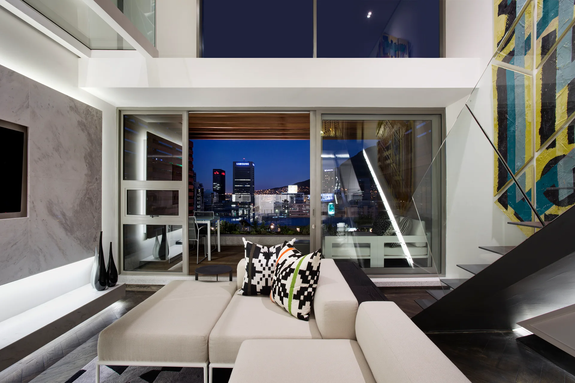 A contemporary double-height living room features a cream sectional sofa, a black open staircase with glass railings, and glass doors opening to a terrace.