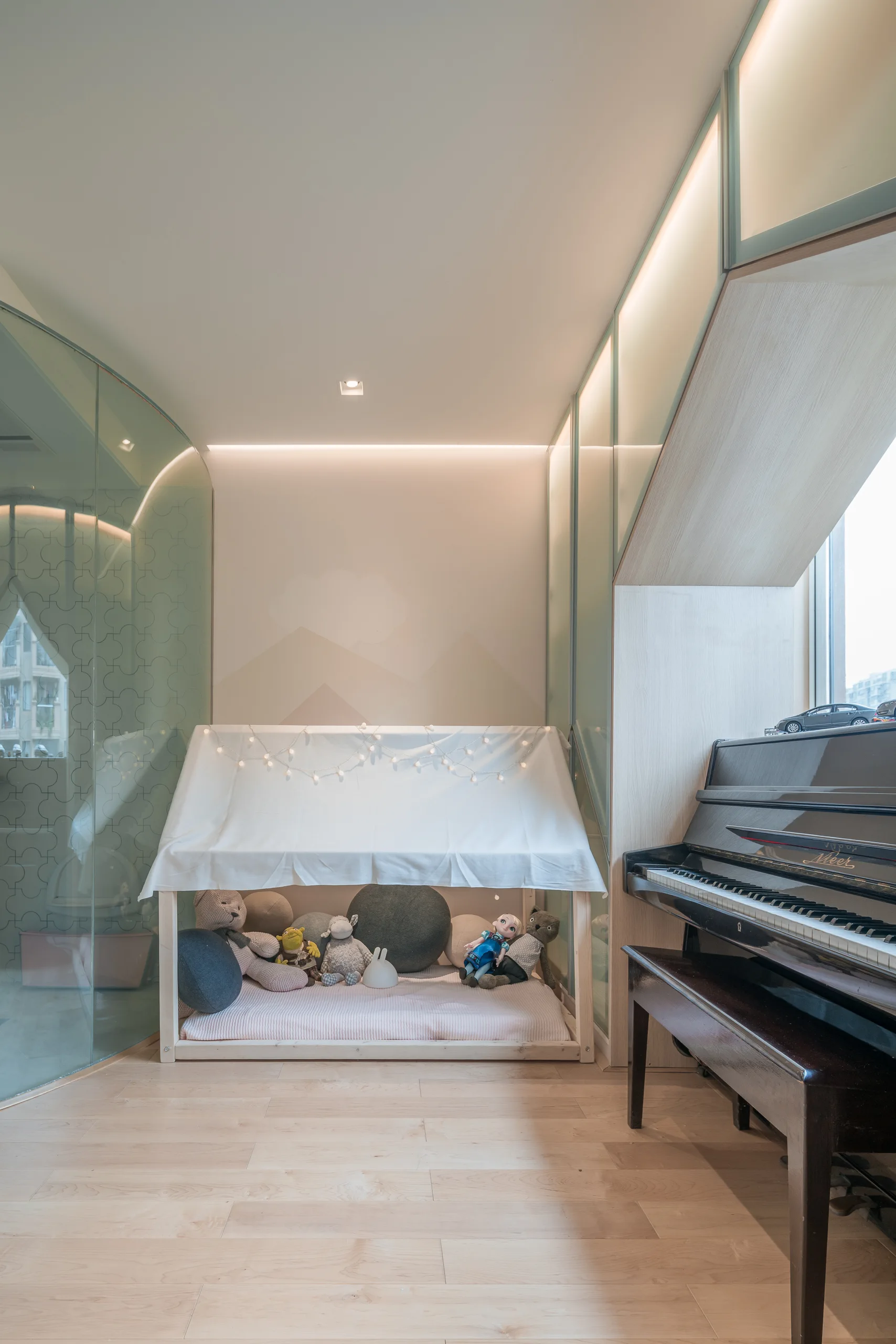 A minimalist children's room features a wooden house-shaped floor bed with string lights and a black upright piano on light wood floors.