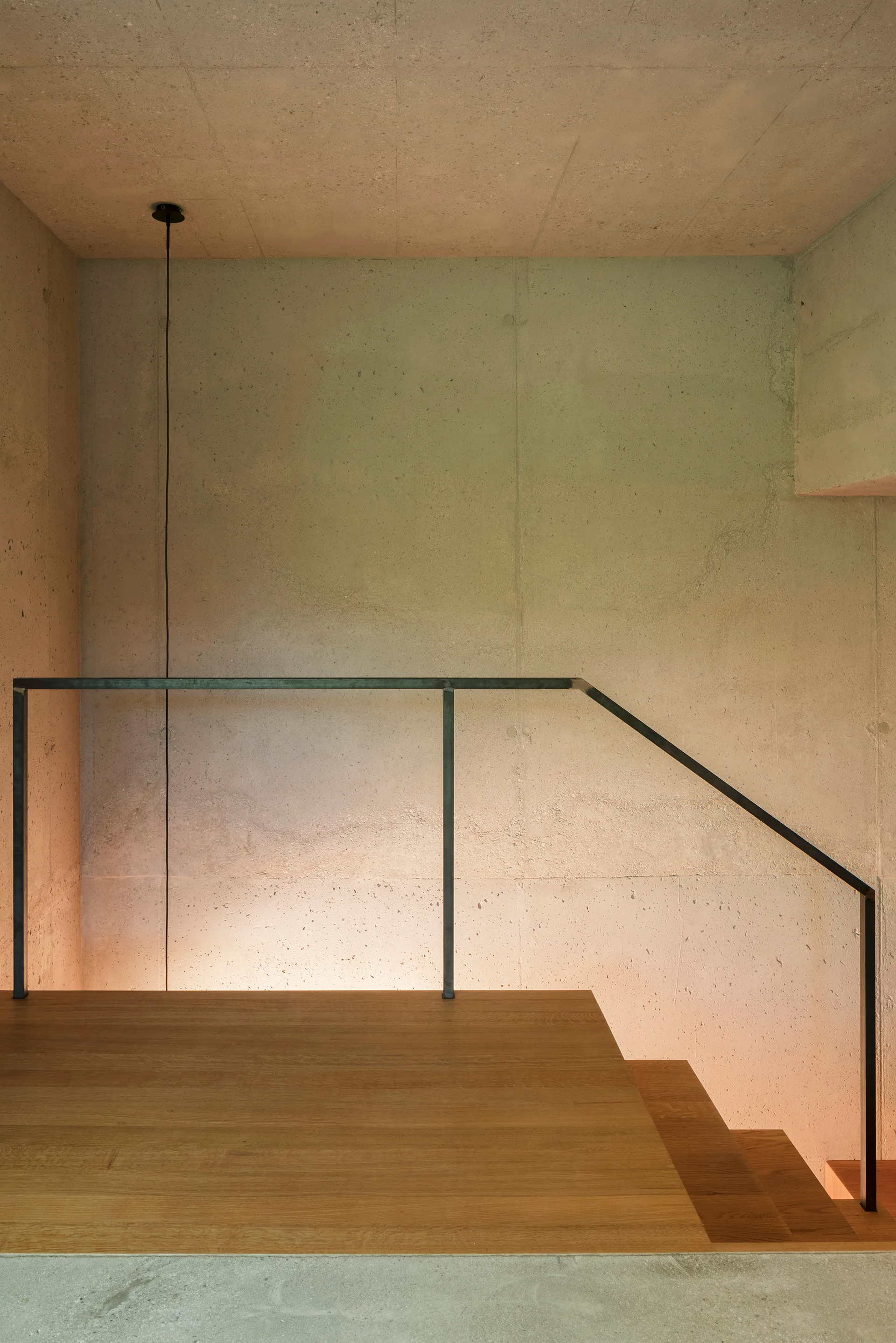 A minimalist staircase landing features warm wood flooring, a thin black metal handrail, and an exposed raw concrete wall.