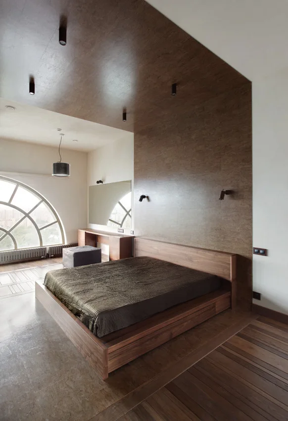 A minimalist bedroom features a wooden platform bed set against a dark wood feature wall that extends onto the ceiling next to a large arched window.