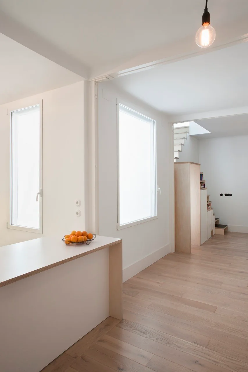 A minimalist white interior features a wood-edged console table, tall windows, light wood flooring, and a staircase in the background.