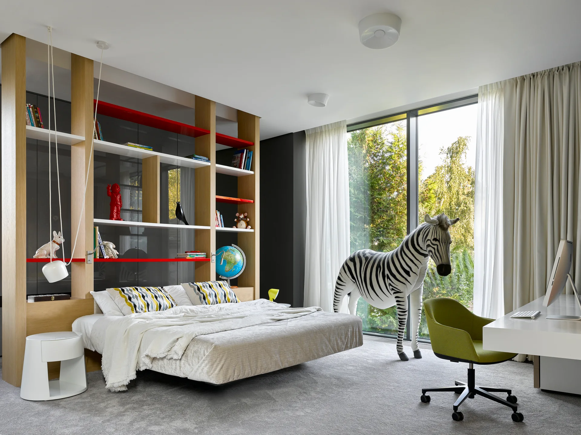 A modern children's bedroom features a low bed in front of a tall wooden shelving unit, a white desk with a green chair, and a large plush zebra.