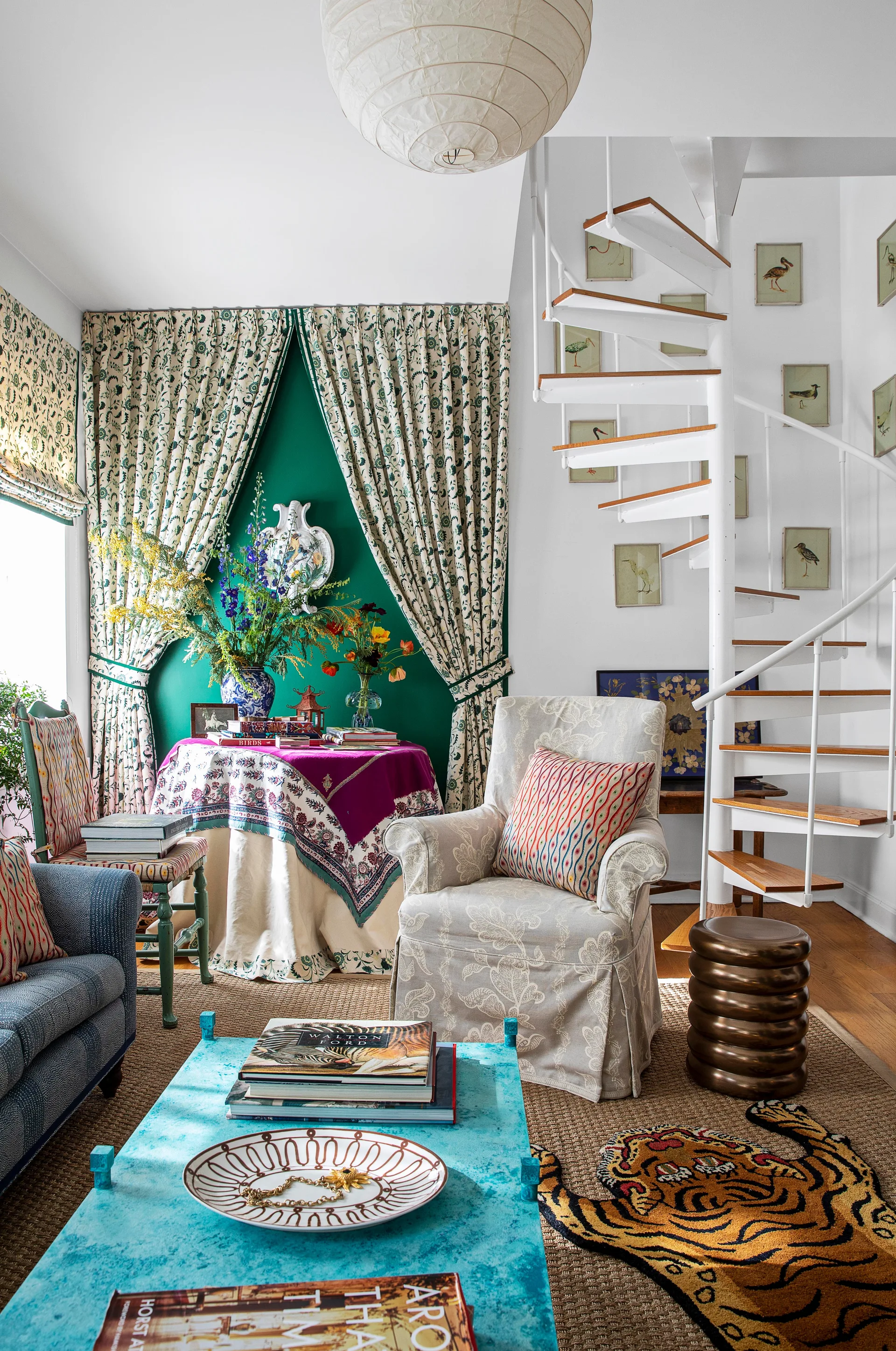 An eclectic living room featuring a white spiral staircase next to a patterned armchair, a turquoise coffee table, and a bold green accent wall.