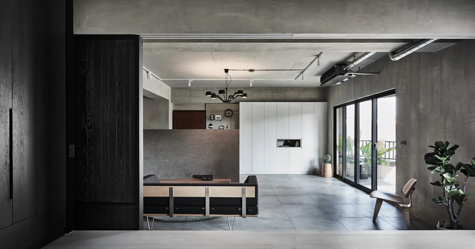 An industrial open-plan living room with exposed concrete walls, a dark sofa, a light wood chair, and white flat-panel storage cabinets.