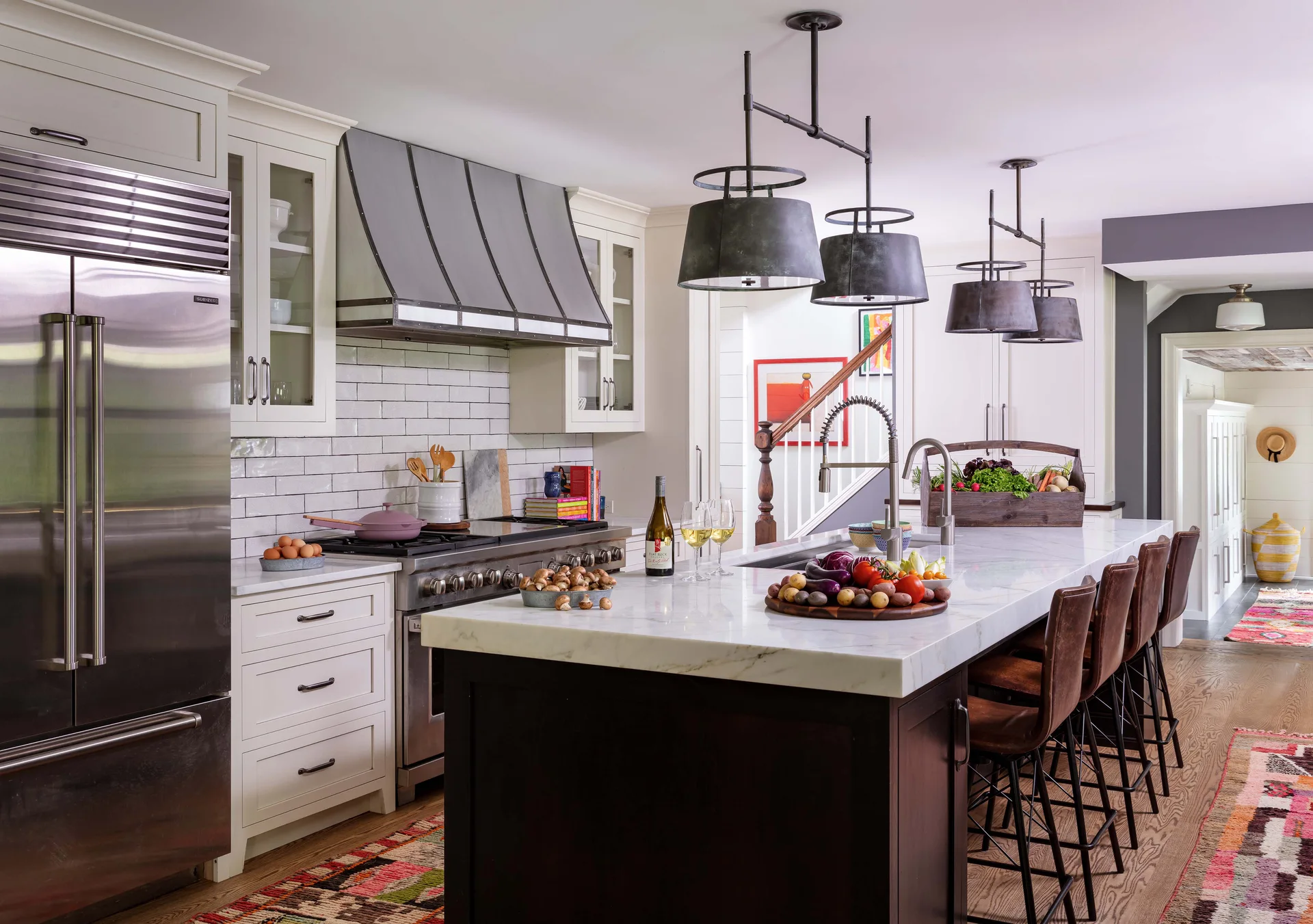 A bright kitchen features a large white marble island with leather bar stools, industrial pendant lighting, and a custom steel range hood.