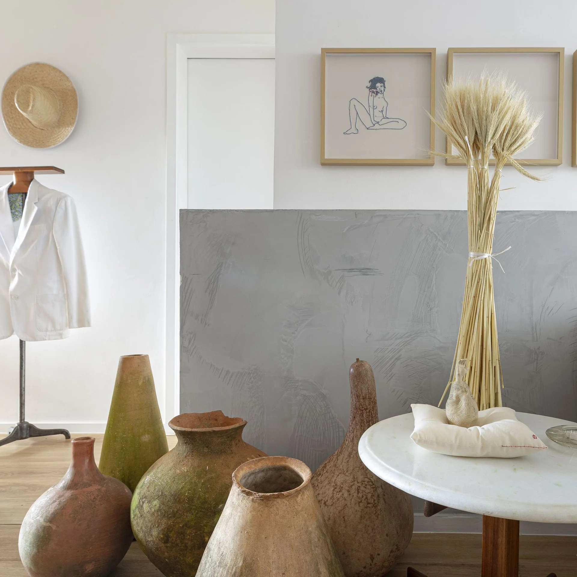 A cluster of large rustic clay pots sits on a wood floor next to a white side table with dried wheat stalks against a textured grey wall panel.
