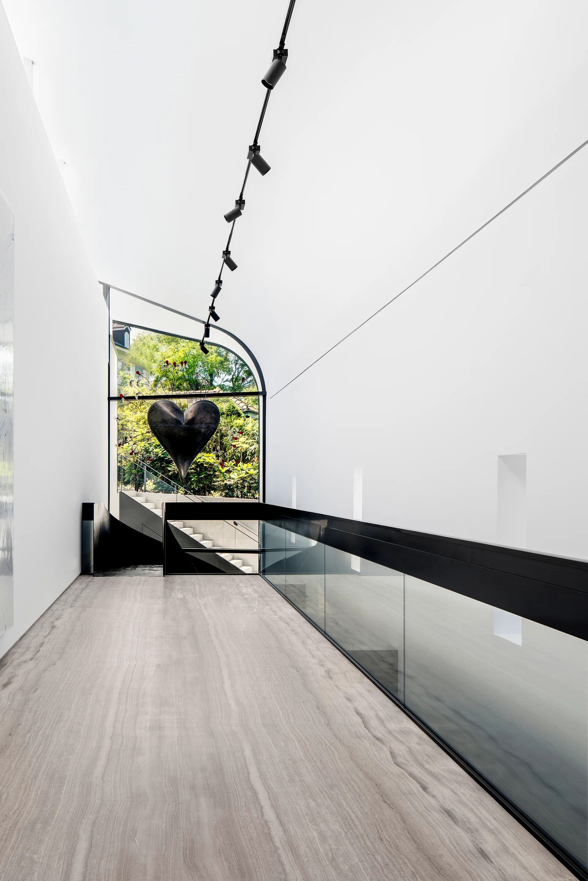 A modern landing features veined grey stone flooring, a glass railing with black trim, and an arched window showing a suspended heart-shaped sculpture.