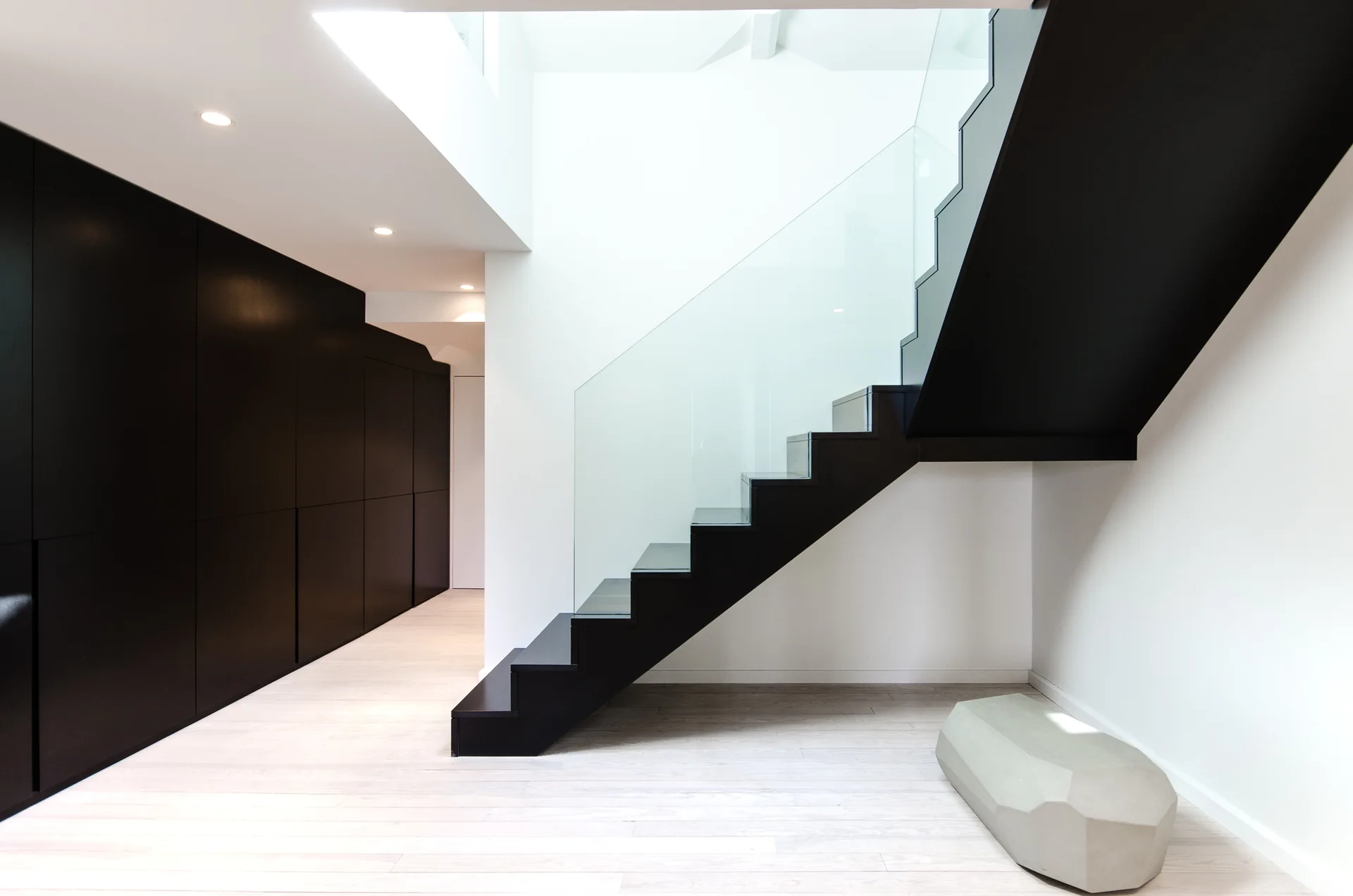 A modern minimalist hallway features a bold black staircase with a clear glass railing opposite matte black built-in storage cabinets.