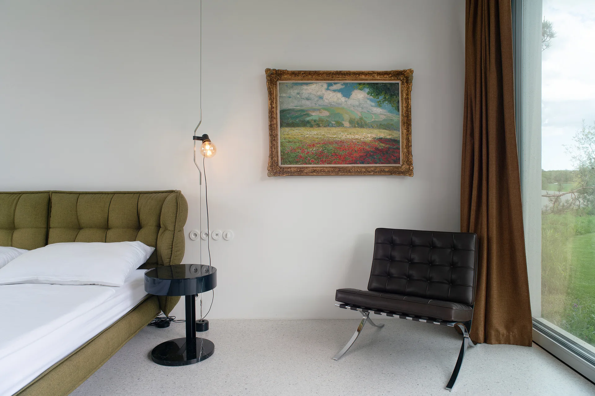 An olive green tufted bed stands next to a black leather armchair and an ornate gold-framed landscape painting in a minimalist white bedroom.