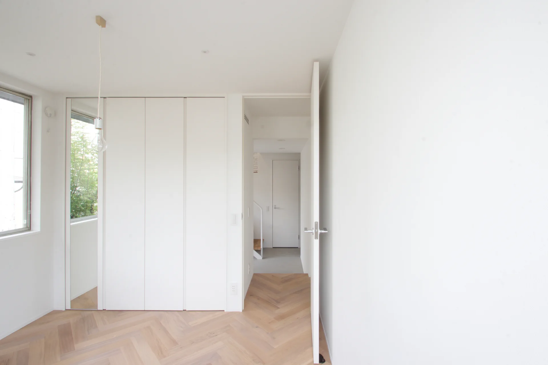 A minimalist white room features light-toned herringbone wood floors, built-in closet panels, and a partially open door leading to a hallway.