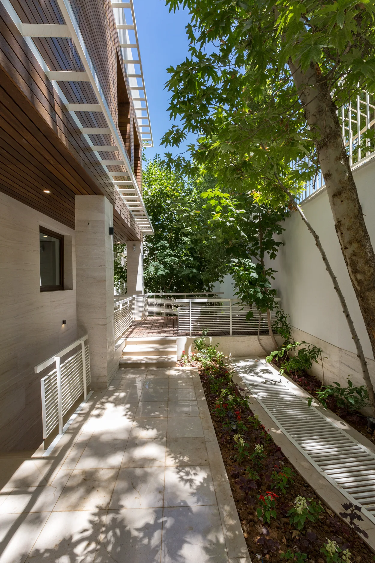 A modern side courtyard features polished stone tile flooring, a narrow garden bed, white metal railings, and a tiered wood deck.