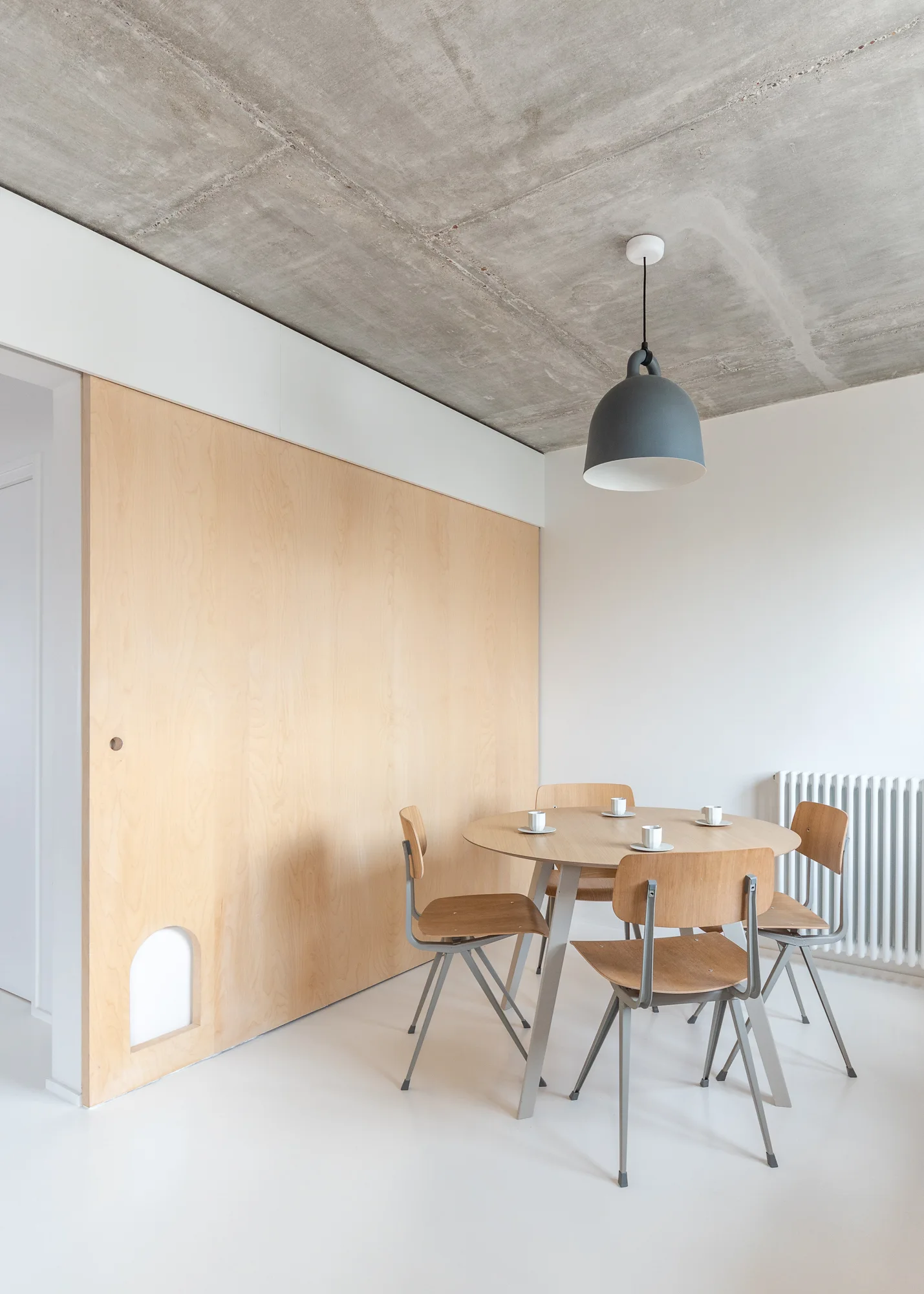 A minimalist dining room features a round wooden table and chairs under a grey pendant light, set against a plywood wall and raw concrete ceiling.