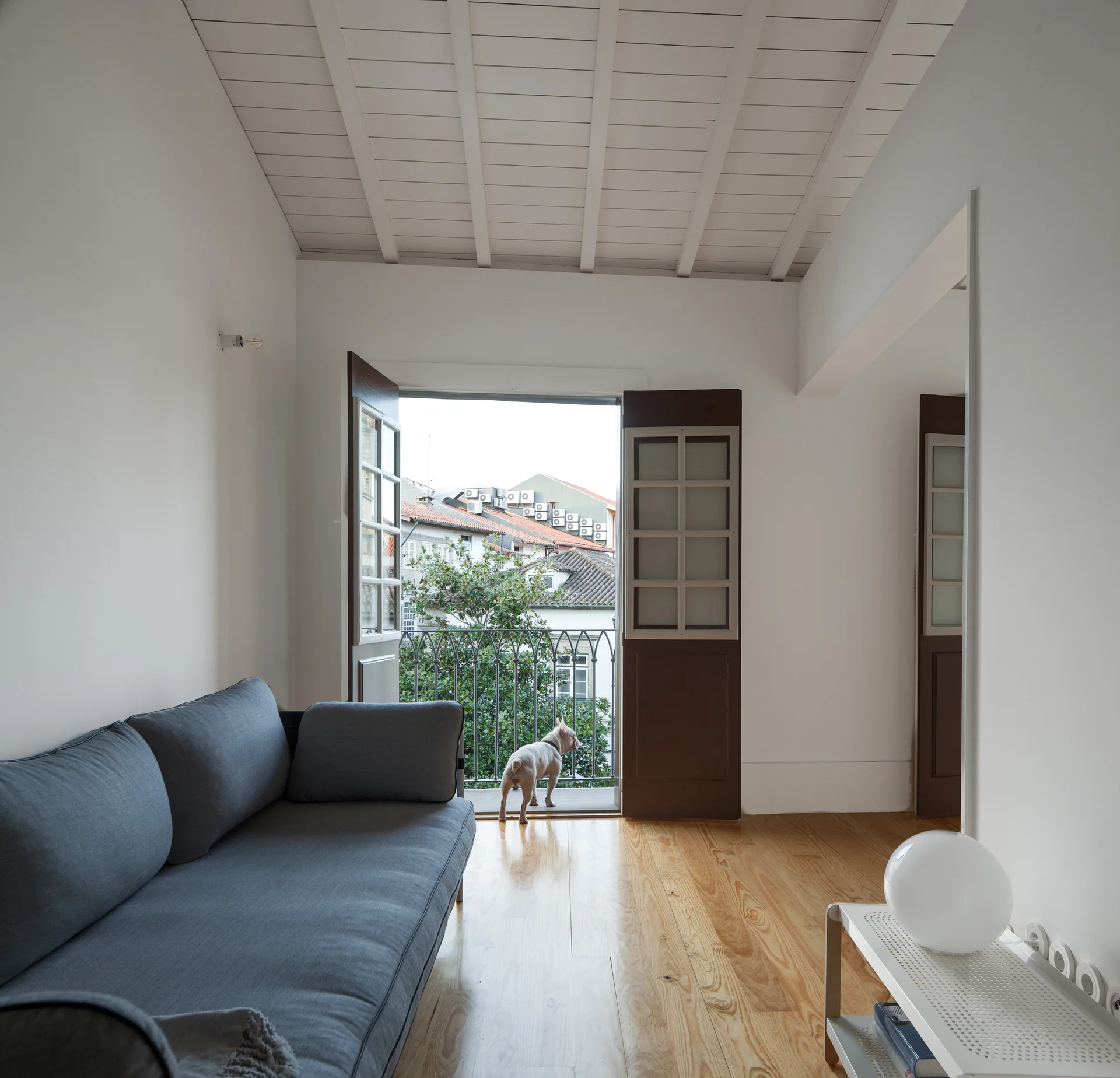 A minimalist living room featuring a grey fabric sofa, light wood floors, a painted slatted ceiling, and open balcony doors leading outside.