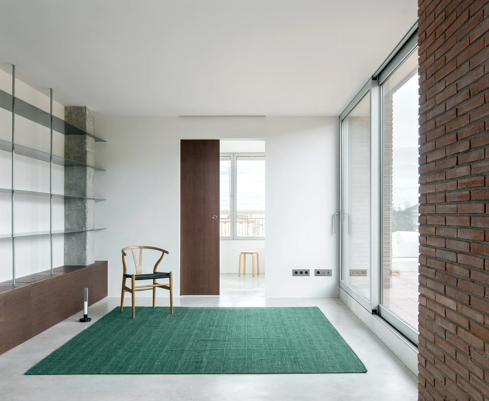 A minimalist room features a wooden wishbone chair on a green rug, next to metal shelving and an exposed red brick wall beside large glass doors.
