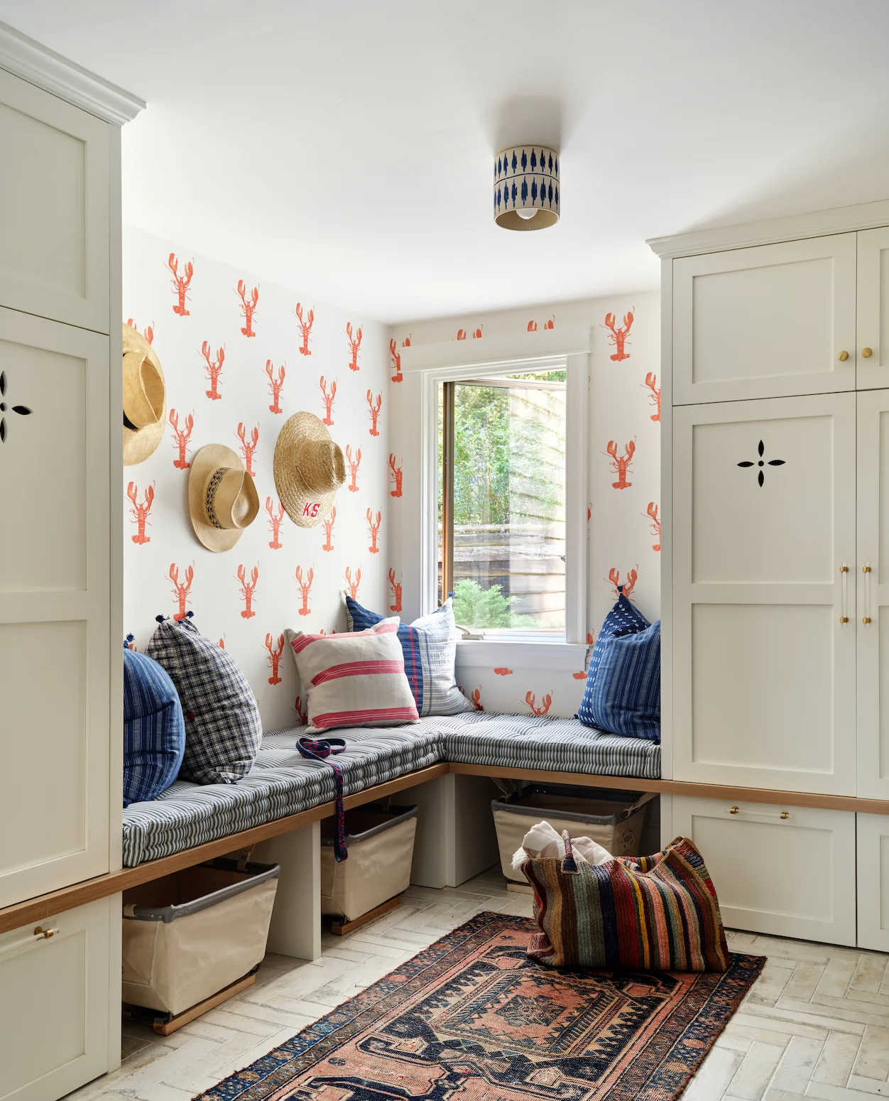 A mudroom features an L-shaped cushioned bench beneath lobster-patterned wallpaper, flanked by tall white storage cabinets.