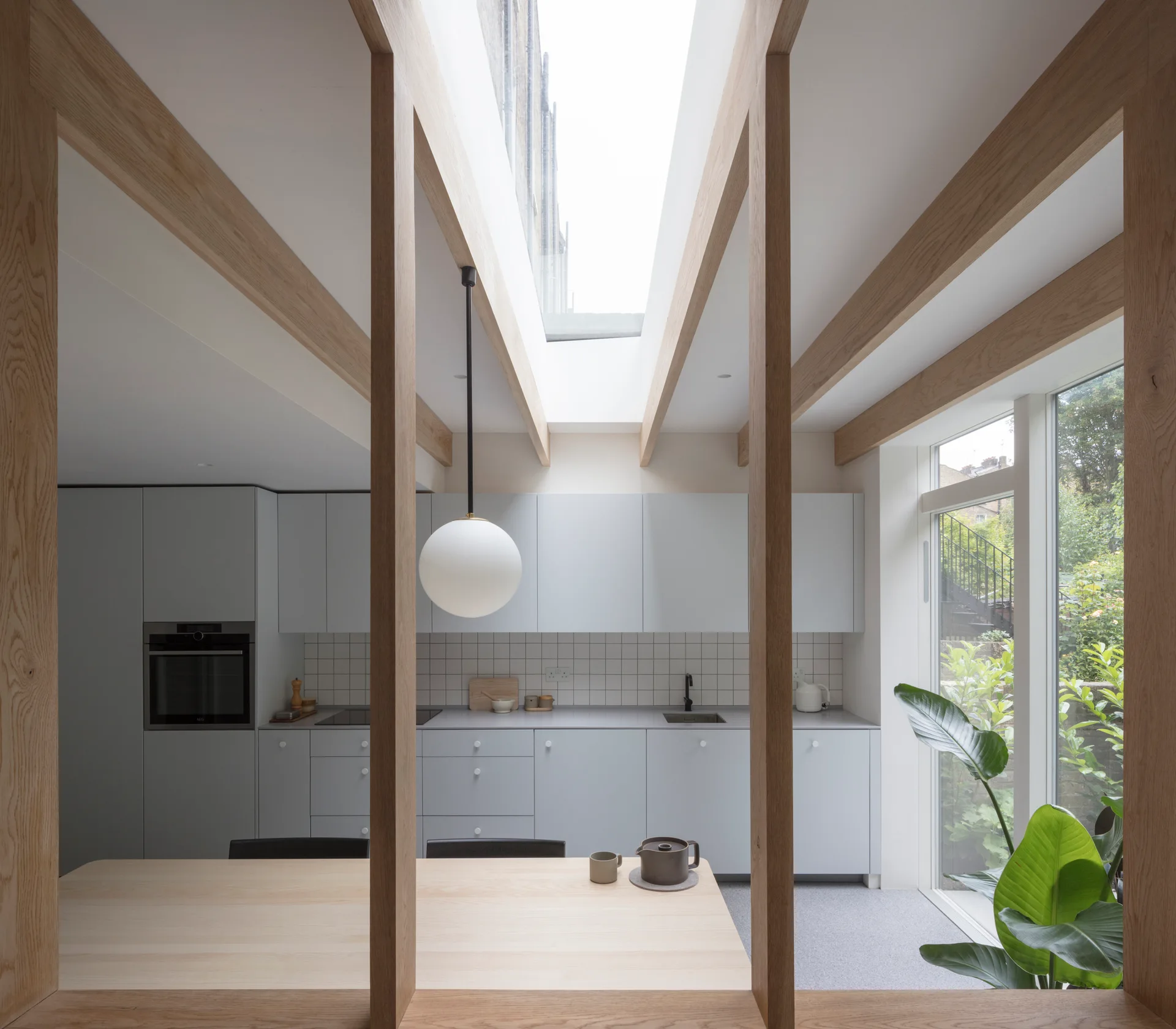 A modern kitchen features light grey cabinets, a square tiled backsplash, exposed wood ceiling beams, a central skylight, and a dining table.