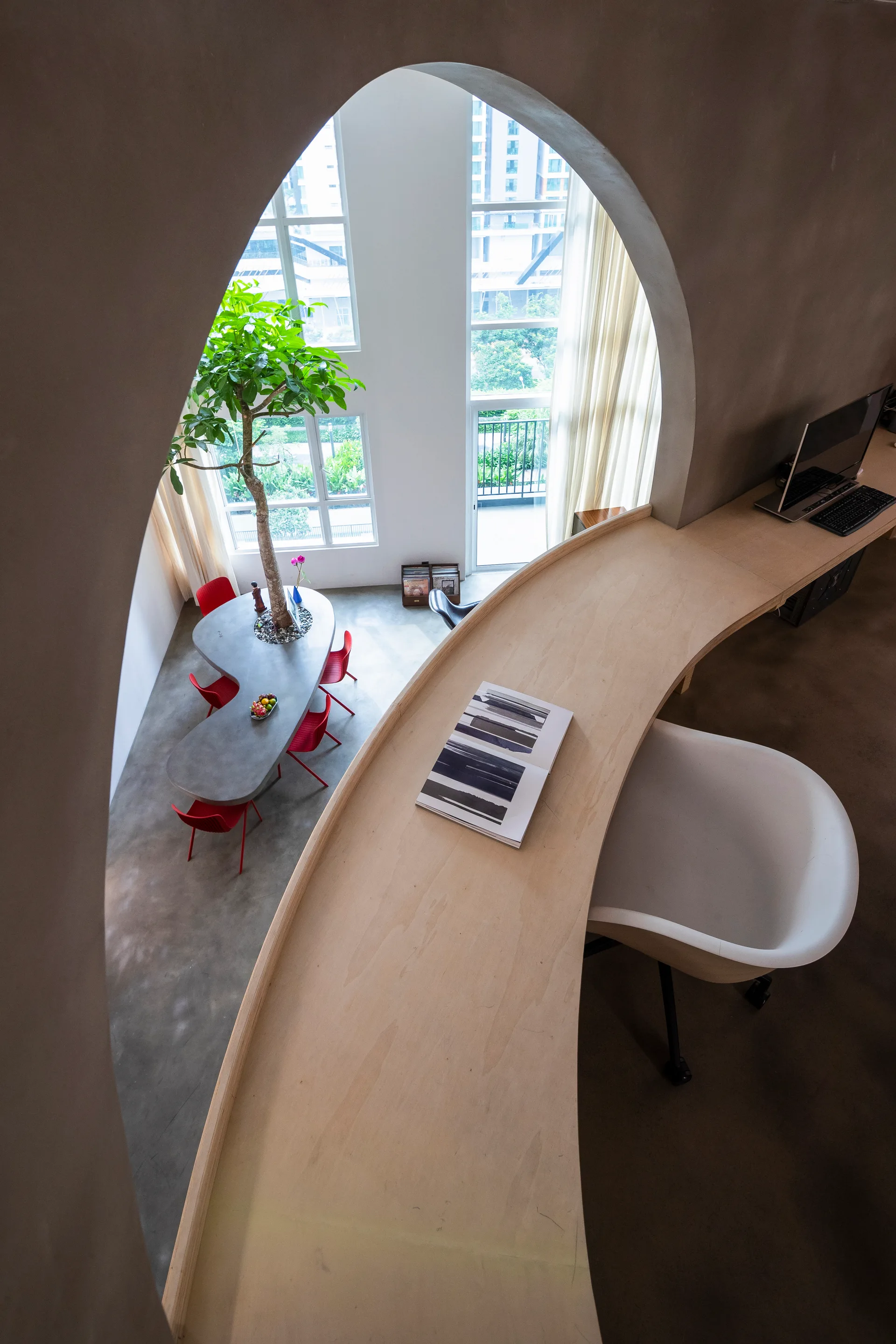 A high-angle view looking through a plaster archway from a mezzanine office with a curved wood desk to a dining area with an indoor tree.