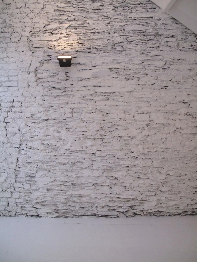 A textured white-painted brick wall features a single black wall sconce shining a beam of light upward.