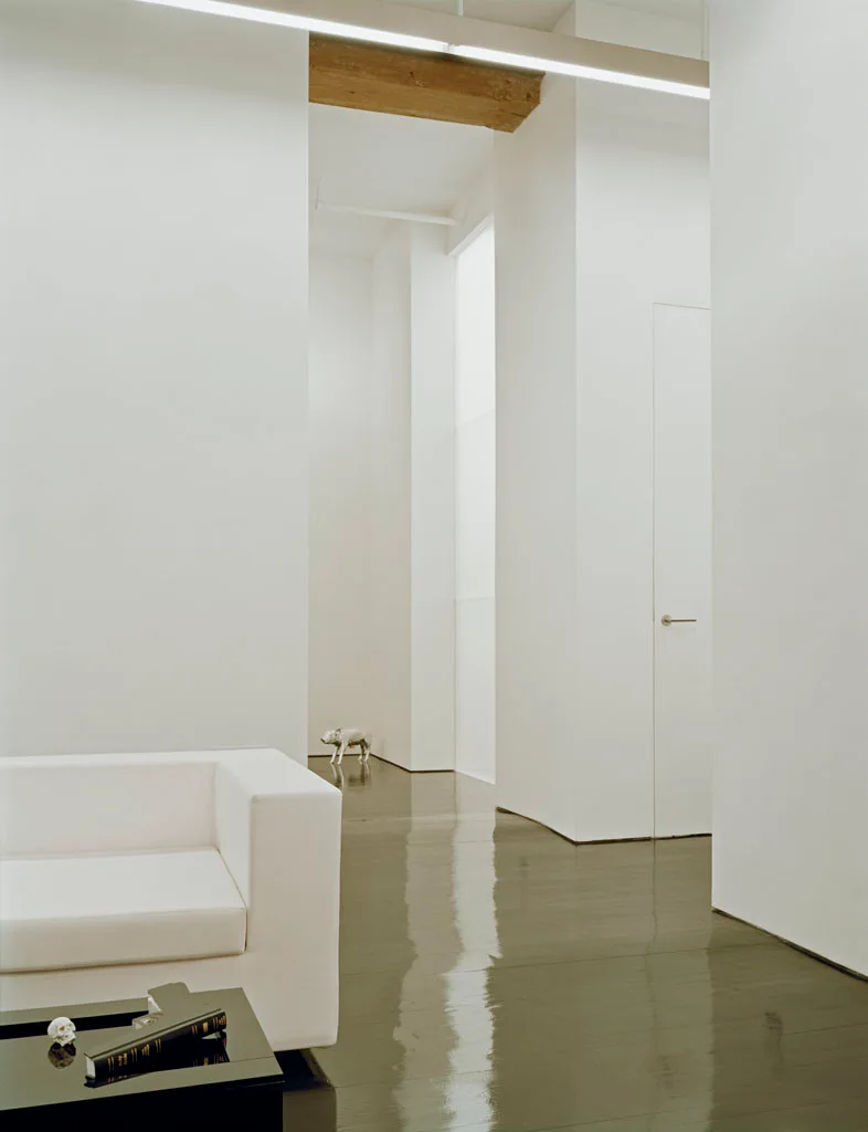 A minimalist living space features a white sofa, high-gloss dark flooring, crisp white walls, and a flush-mounted door.
