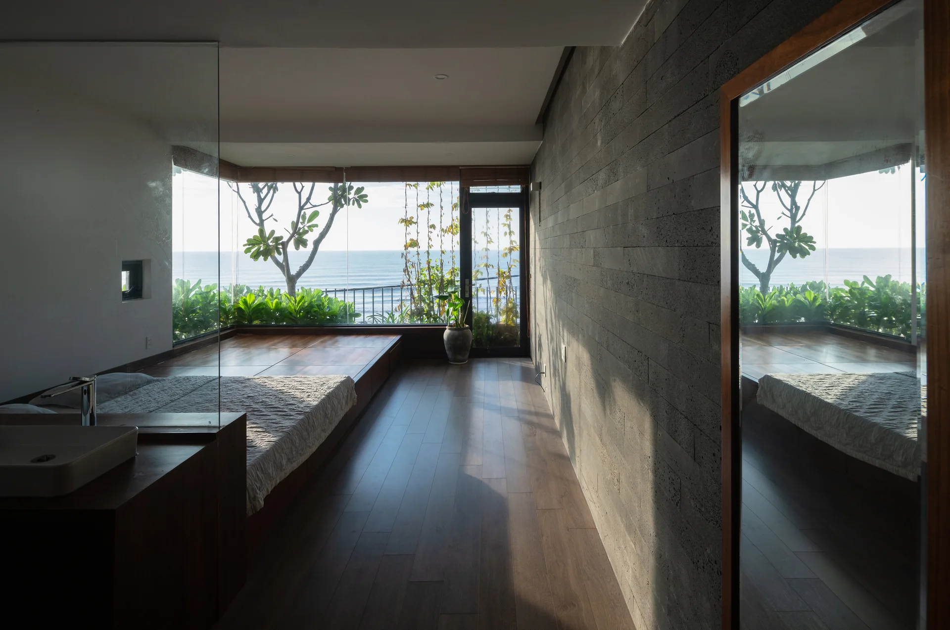 A minimalist bedroom features a raised wooden bed platform, a textured grey stone wall, and a large window looking out at the ocean.