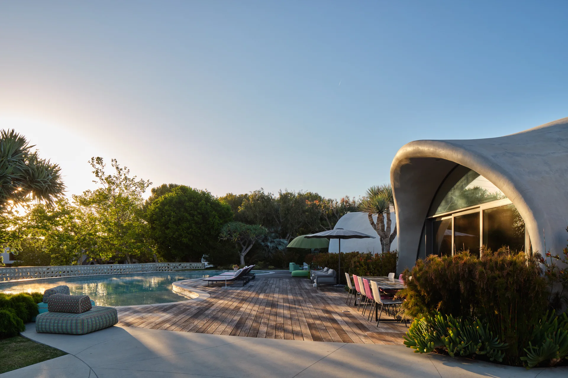 A curved swimming pool sits adjacent to a weathered wood deck and a unique shell-shaped concrete dome structure with outdoor dining and lounge furniture.
